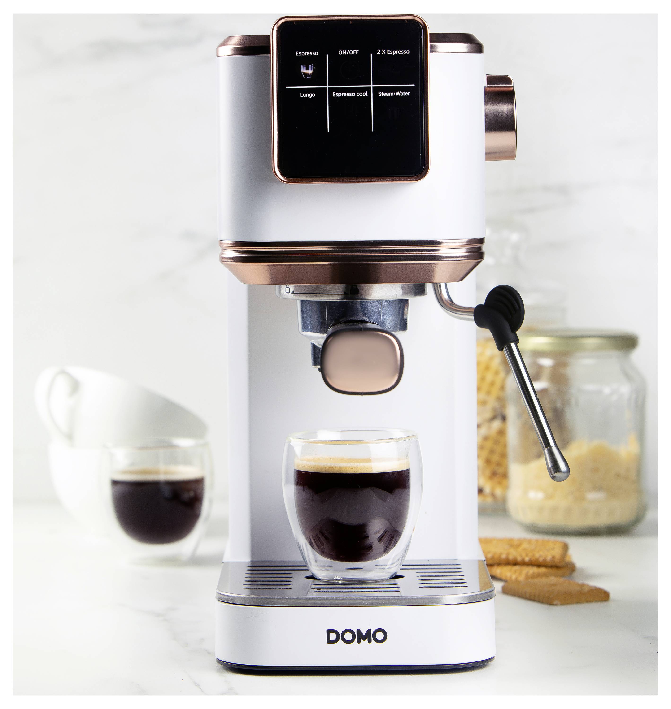 Kávovar na espresso připravující kávu do skleněného šálku, s připojeným napěňovačem mléka. V pozadí šálky a skleněná dóza na sušenky.