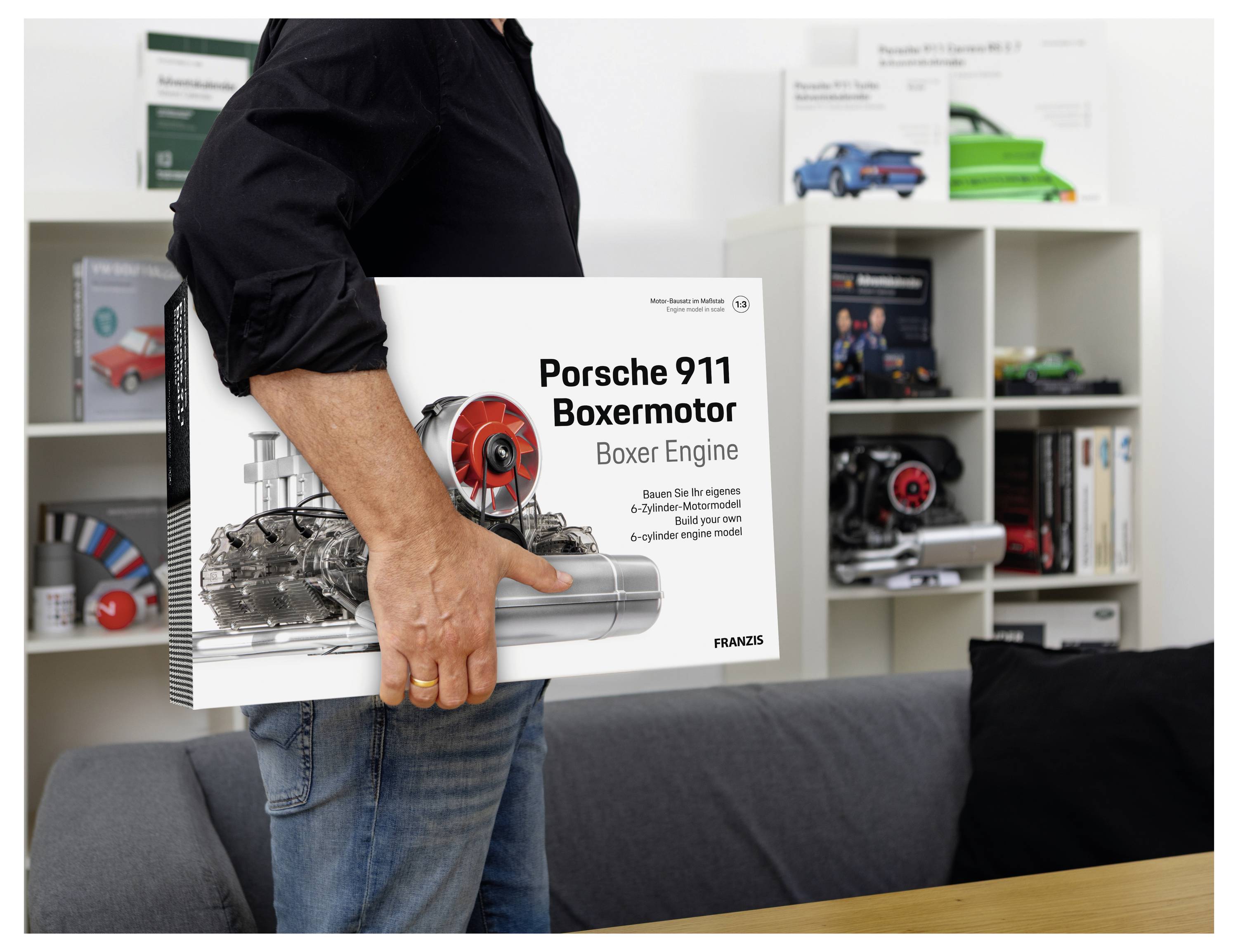 Osoba držící knihu 'Porsche 911 Boxermotor', ukazující diagram motoru, s policemi modelů aut a knih v pozadí.