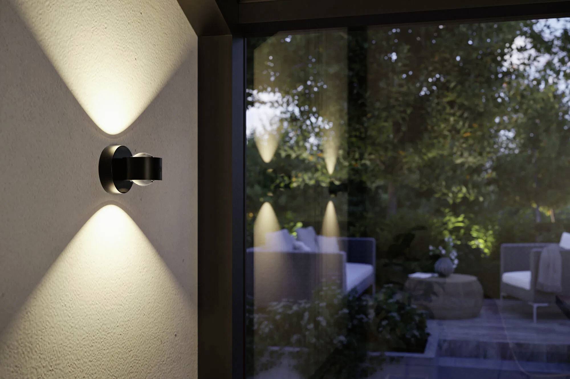 Nástěnná lampa osvětlující terasu za soumraku, s venkovním posezením viditelným skrze skleněné dveře, obklopená zelení a tlumeným osvětlením.