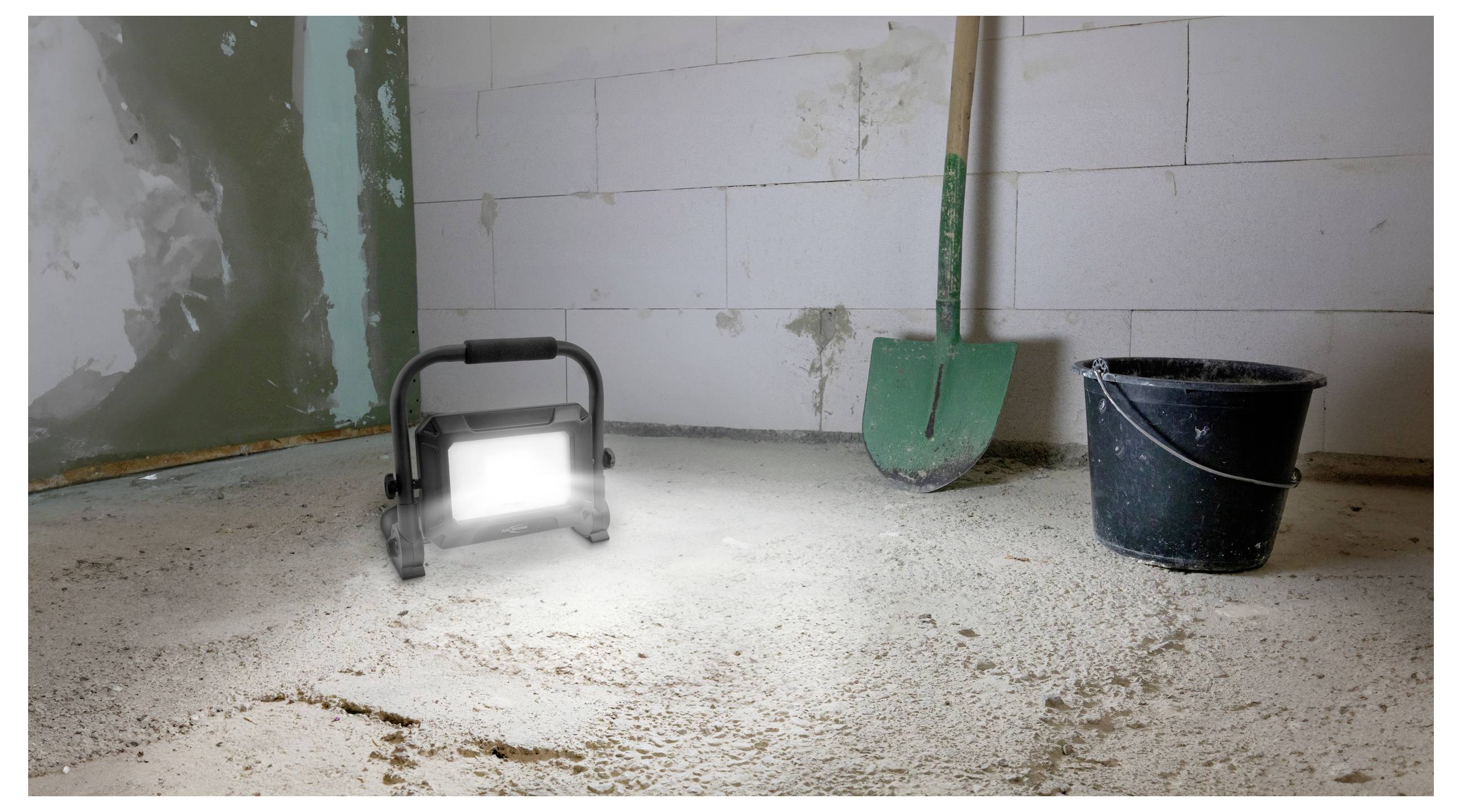 Matně osvětlená místnost ve stadiu rekonstrukce obsahuje zářící pracovní světlo na podlaze, zelené lopaty opřené o zeď a černý kbelík poblíž.