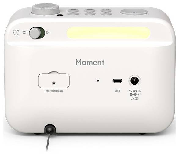 Bílé elektronické zařízení označené nápisem 'Moment' má vypínač, záložní alarm, USB port a napájecí vstup s ovládacím voličem na horní straně.
