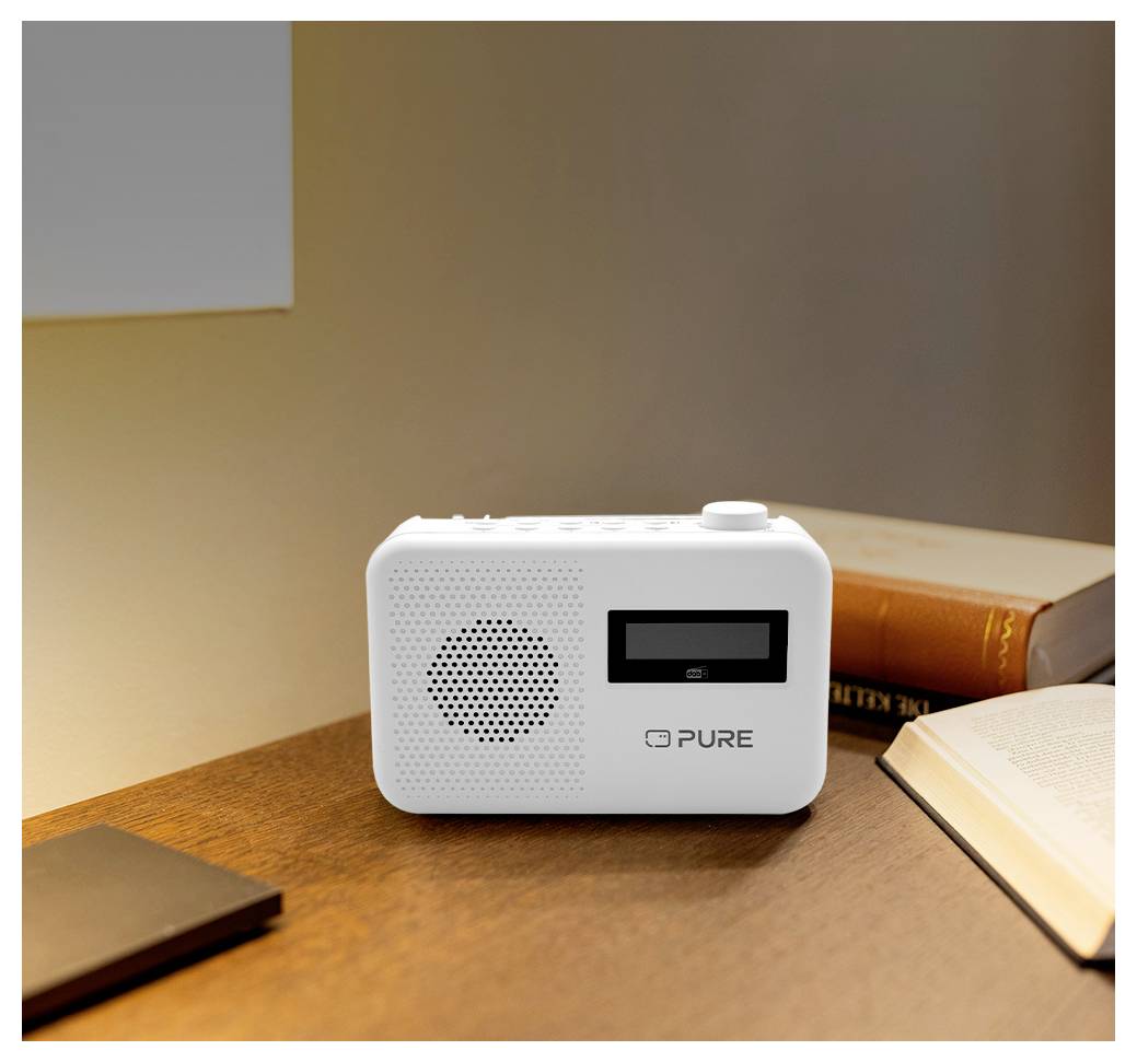 Malé bílé rádio s displejem a reproduktorem leží na dřevěném stole vedle knih, což naznačuje prostor pro čtení nebo studium.