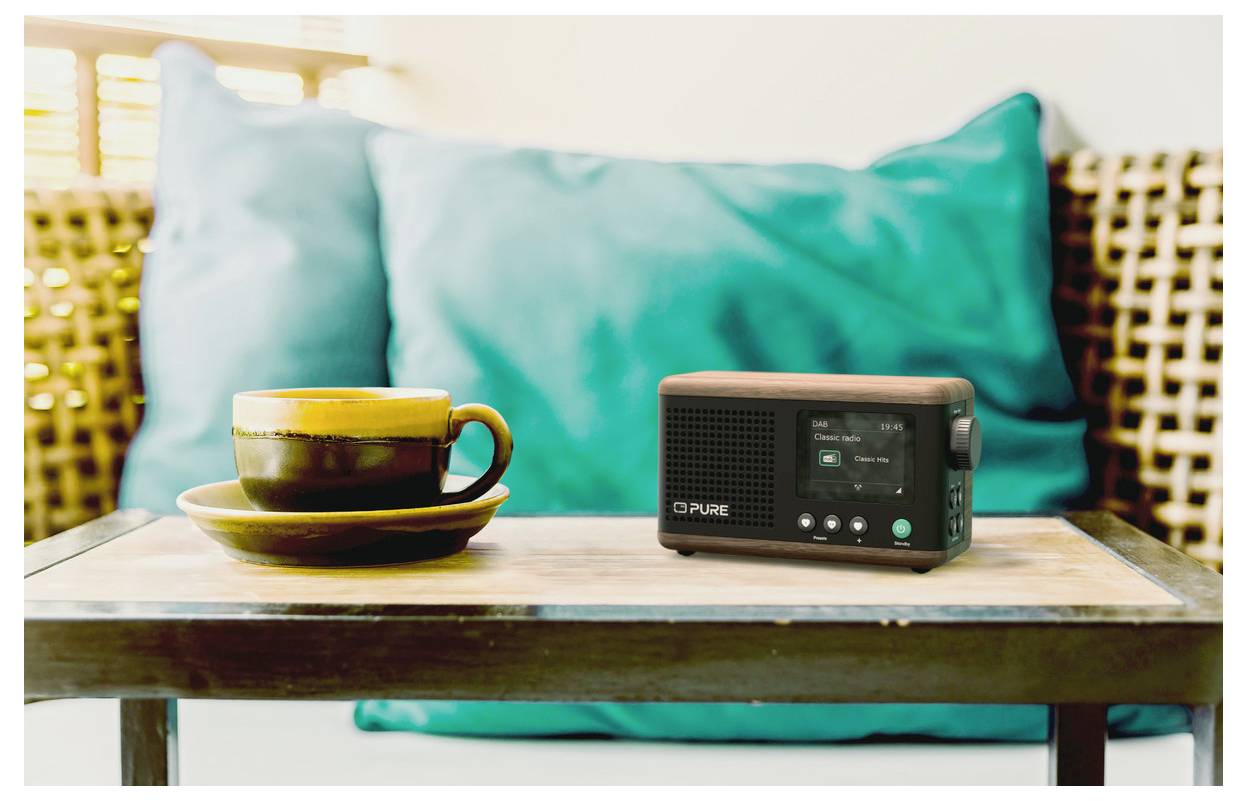 Hrnek kávy a malé rádio leží na stole, s rozmazaným modrým polštářem a tkaným opěradlem v pozadí, což vytváří útulnou atmosféru.