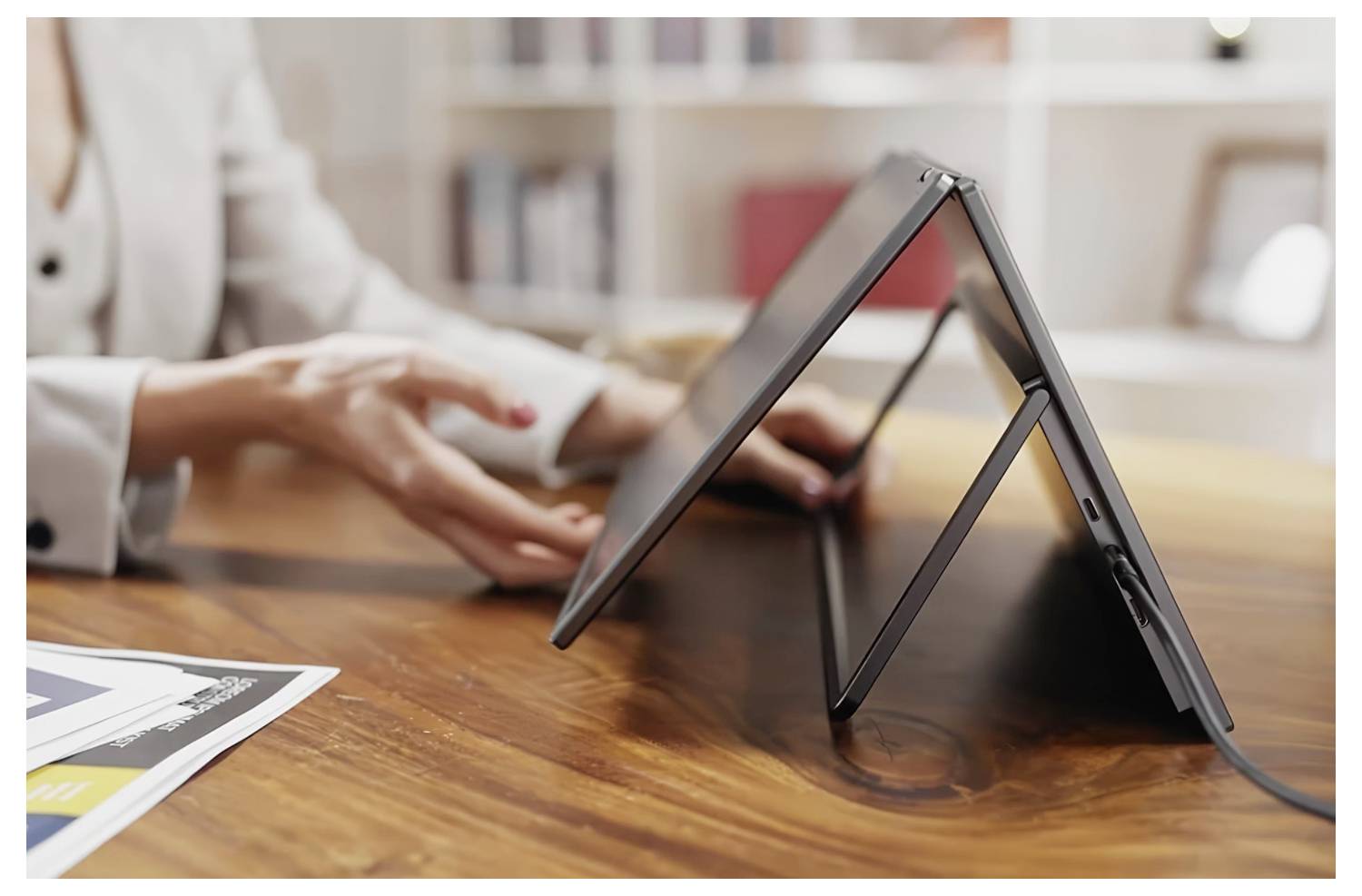Osoba předvádí notebook v módu stanu na dřevěném stole, přičemž zdůrazňuje jeho flexibilní design pro uživatelské pohodlí v různých prostředích.