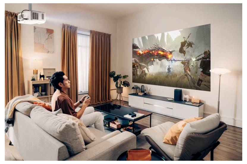 Osoba sedí na gauči a hraje videohru na velkém projektoru, který zobrazuje fantastickou bitevní scénu v moderním obývacím pokoji.