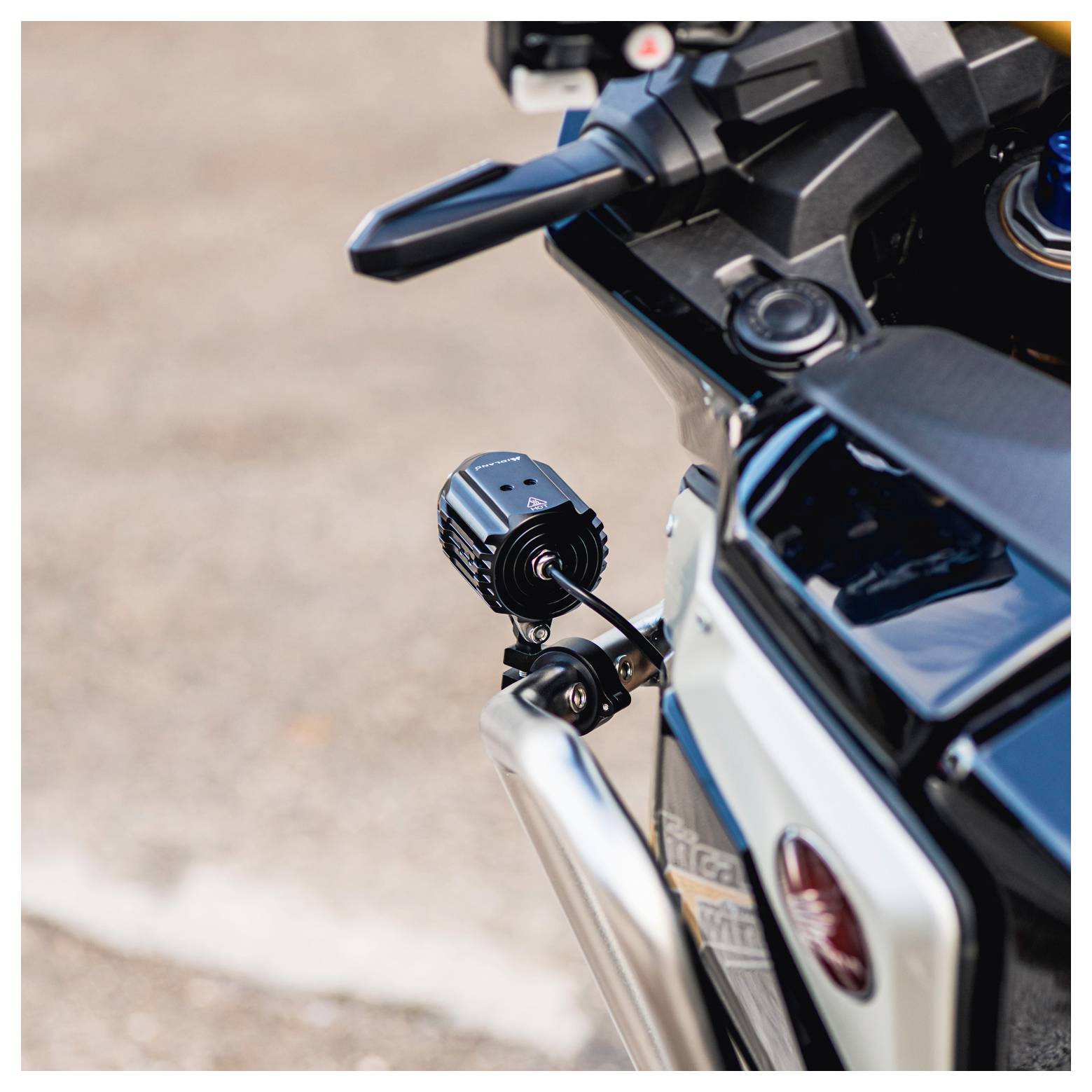 Detailní záběr motocyklové řídítka s připevněnou malou černou kamerou, zachycený z předního pohledu. Pozadí je rozmazaná silnice.