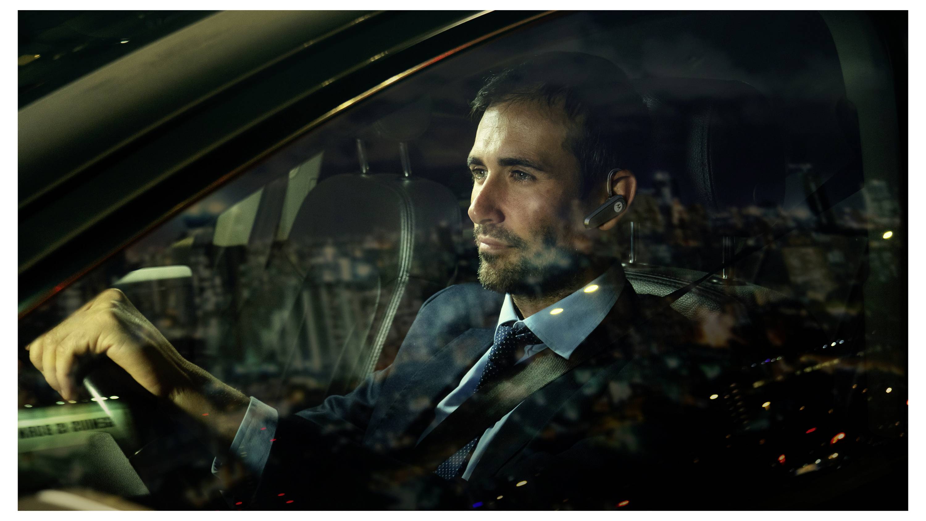 Muž v obleku řídí auto v noci v městské zástavbě, světla města se odrážejí na okně.
