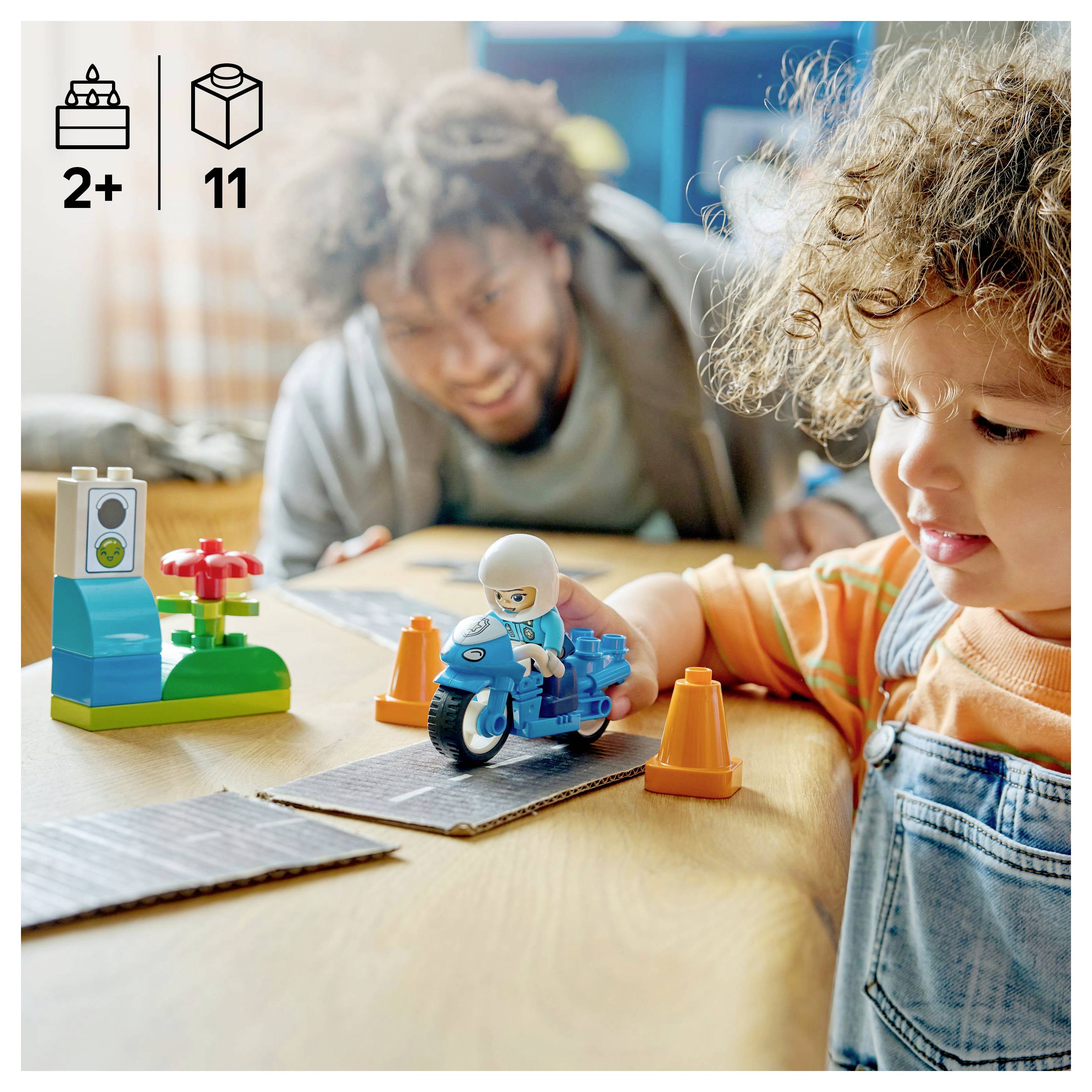 Dítě si hraje s hračkou motocyklu na silniční dráze, v pozadí je dospělý. Na stole jsou stavebnicové kostky a květiny.