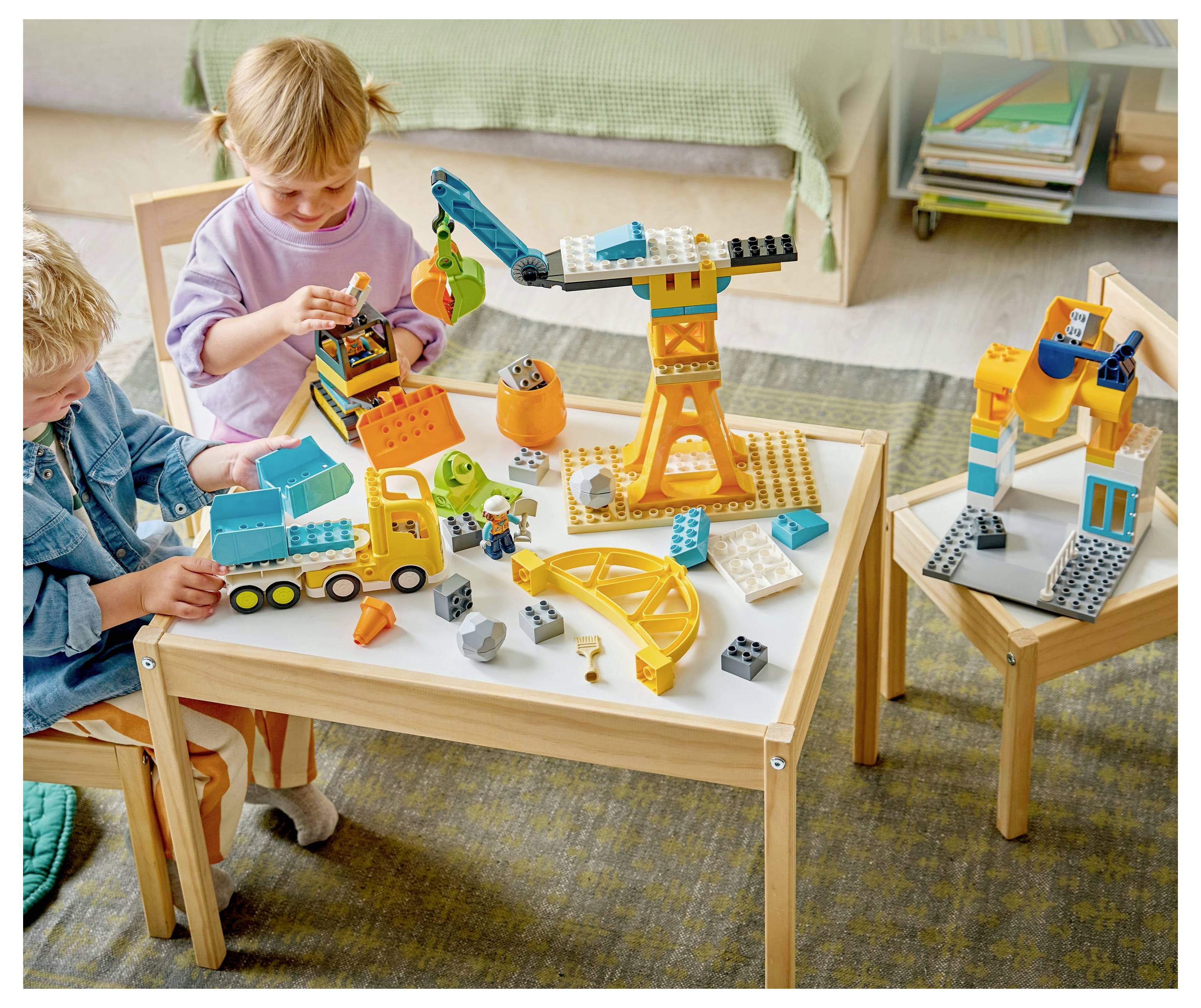 Děti si hrají s barevnými stavebními kostkami u malého stolu, konstruují scénu, která zahrnuje jeřáb a další vozidla.