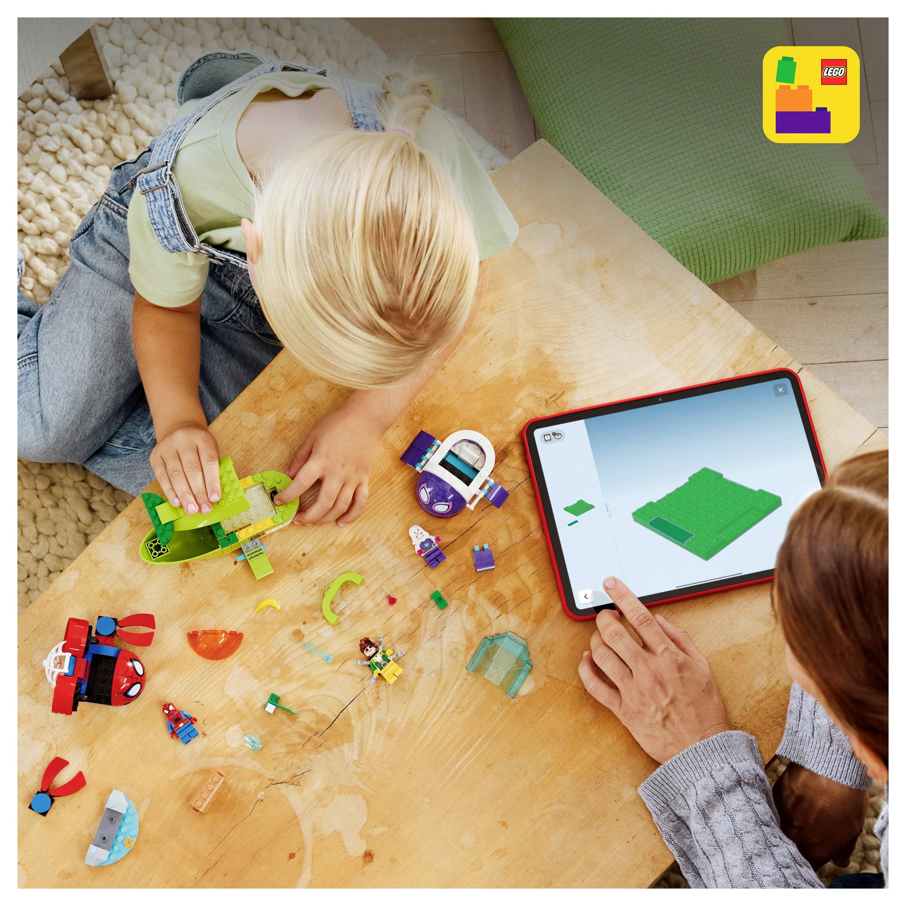 Dítě skládá díly LEGO na stole, přičemž postupuje podle návodu zobrazeného na tabletu. Dospělý pomáhá a ukazuje spolupráci při hře a učení.
