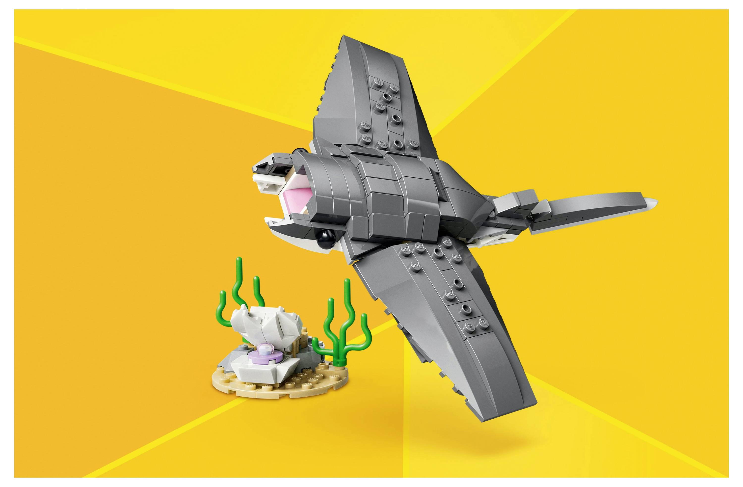 Šedý LEGO model rejnoka s růžovou tlamou plave nad písčitým terénem s zelenou mořskou trávou na žlutém pozadí.