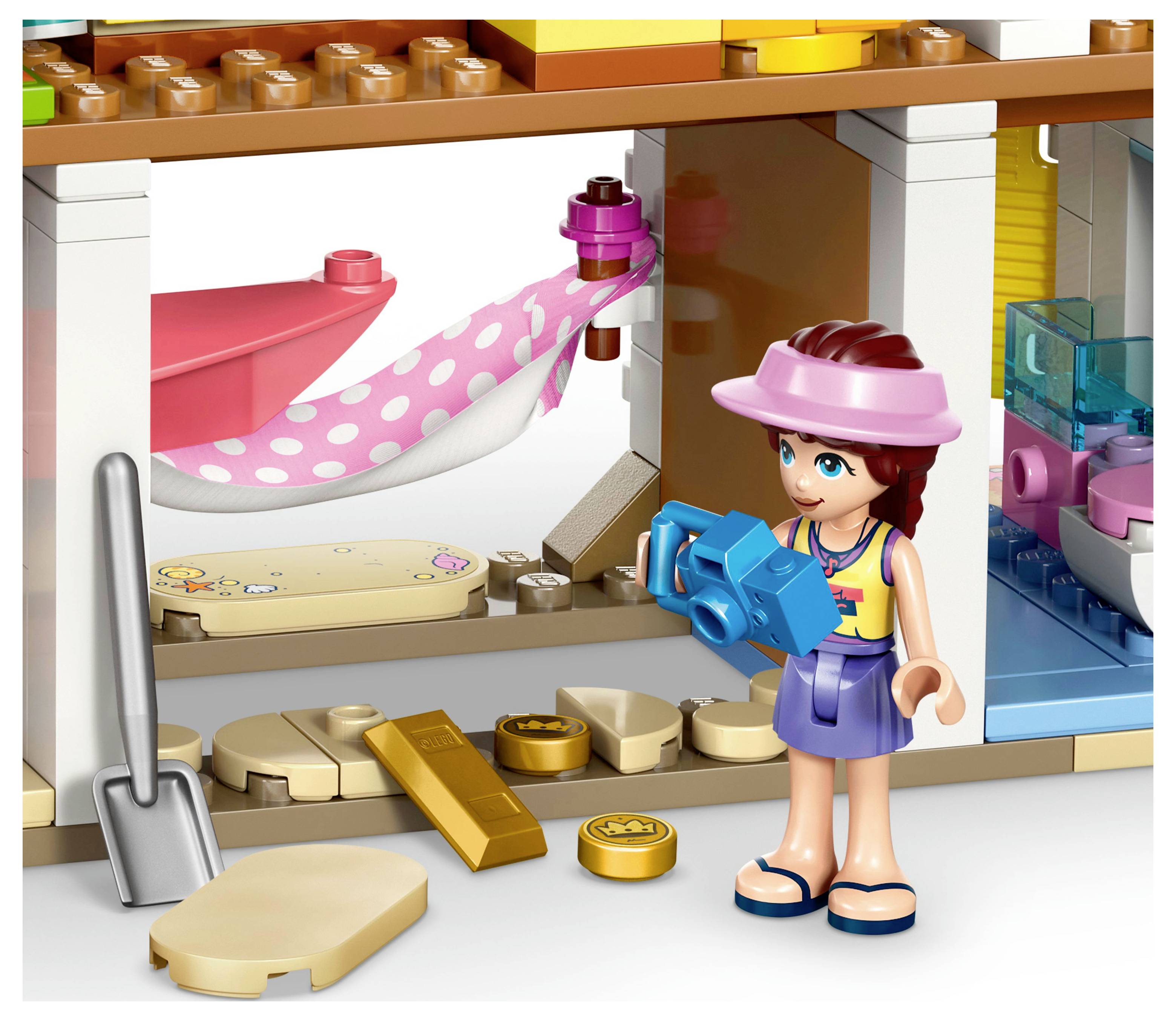 Hračkářská postava s fotoaparátem stojí uvnitř barevného Lego domku. Nachází se zde růžová houpací síť, lopata a rozházené hračkové mince.