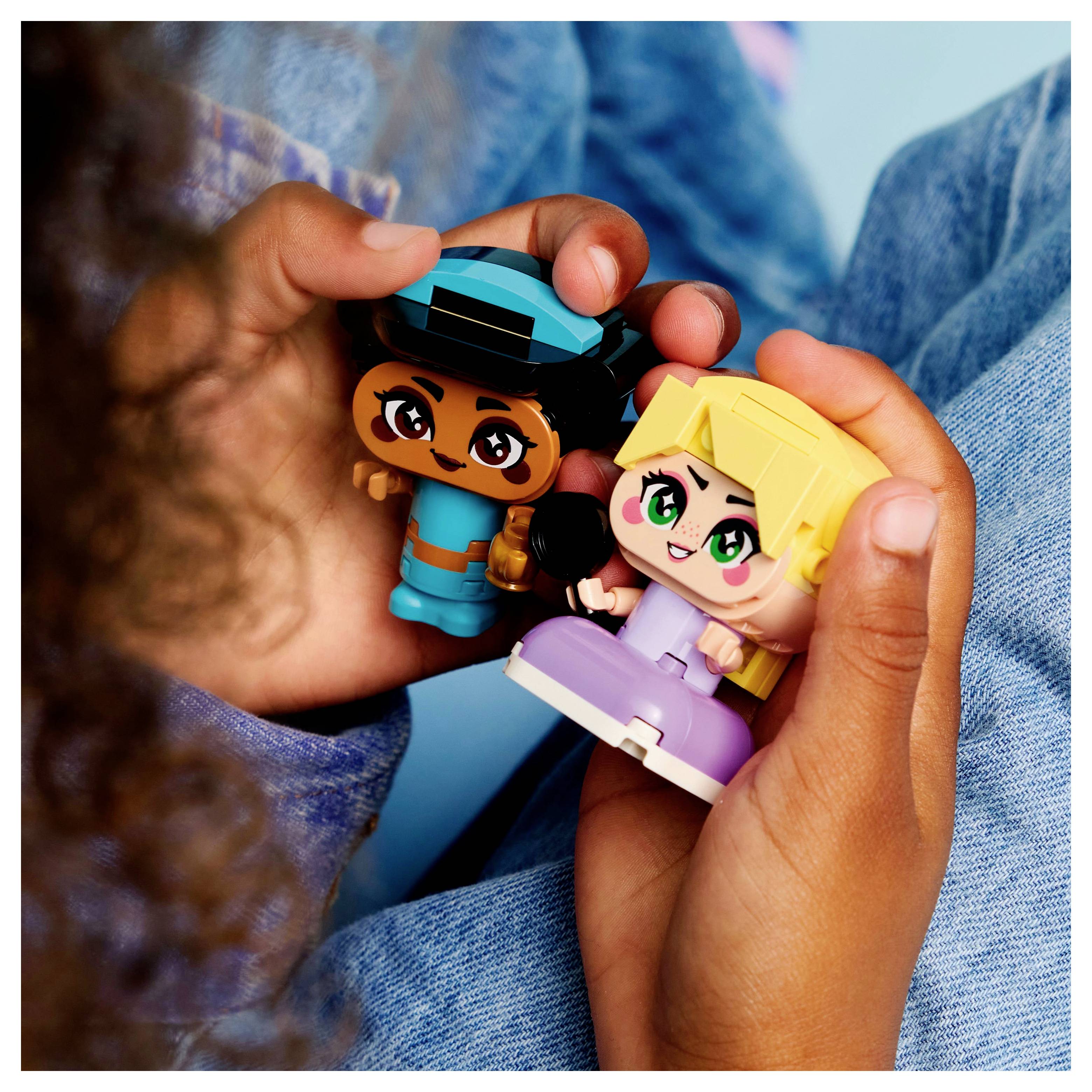 Dvě děti drží barevné hračky, jedna v modrém oblečení s čepicí a druhá ve světle fialovém oblečení, sedící na modré ploše.