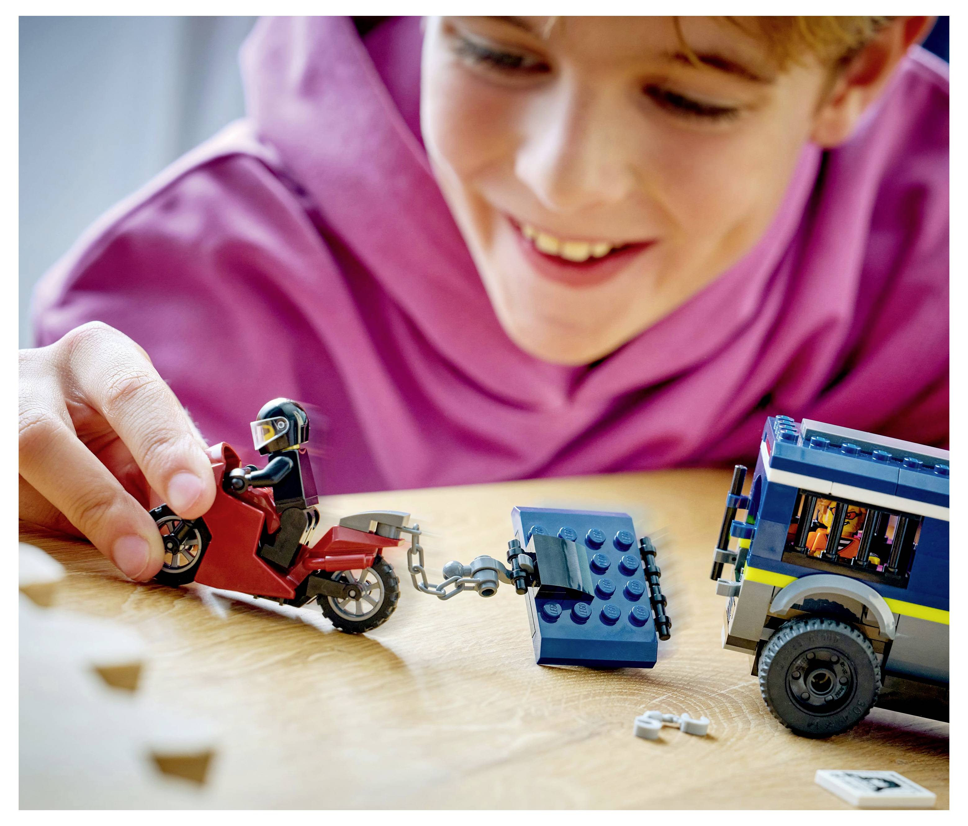 Dítě v růžové mikině si hraje s LEGO hračkami a manévruje červenou motorkou a modrým odtahovým vozem na dřevěném povrchu.