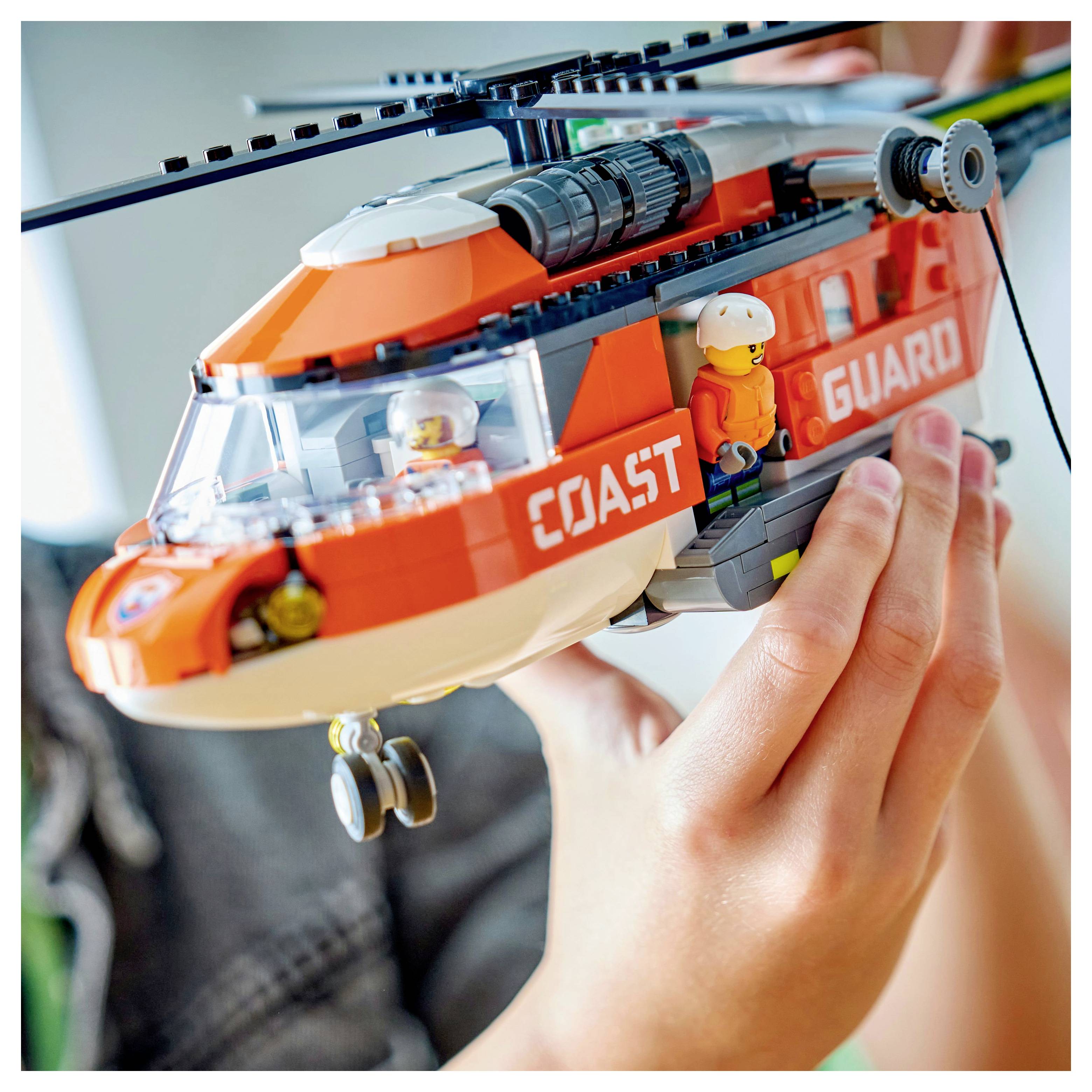 Osoba drží model vrtulníku Pobřežní stráže od LEGO. Vrtulník je oranžový s textem "Pobřežní stráž" a uvnitř obsahuje malé figurky.