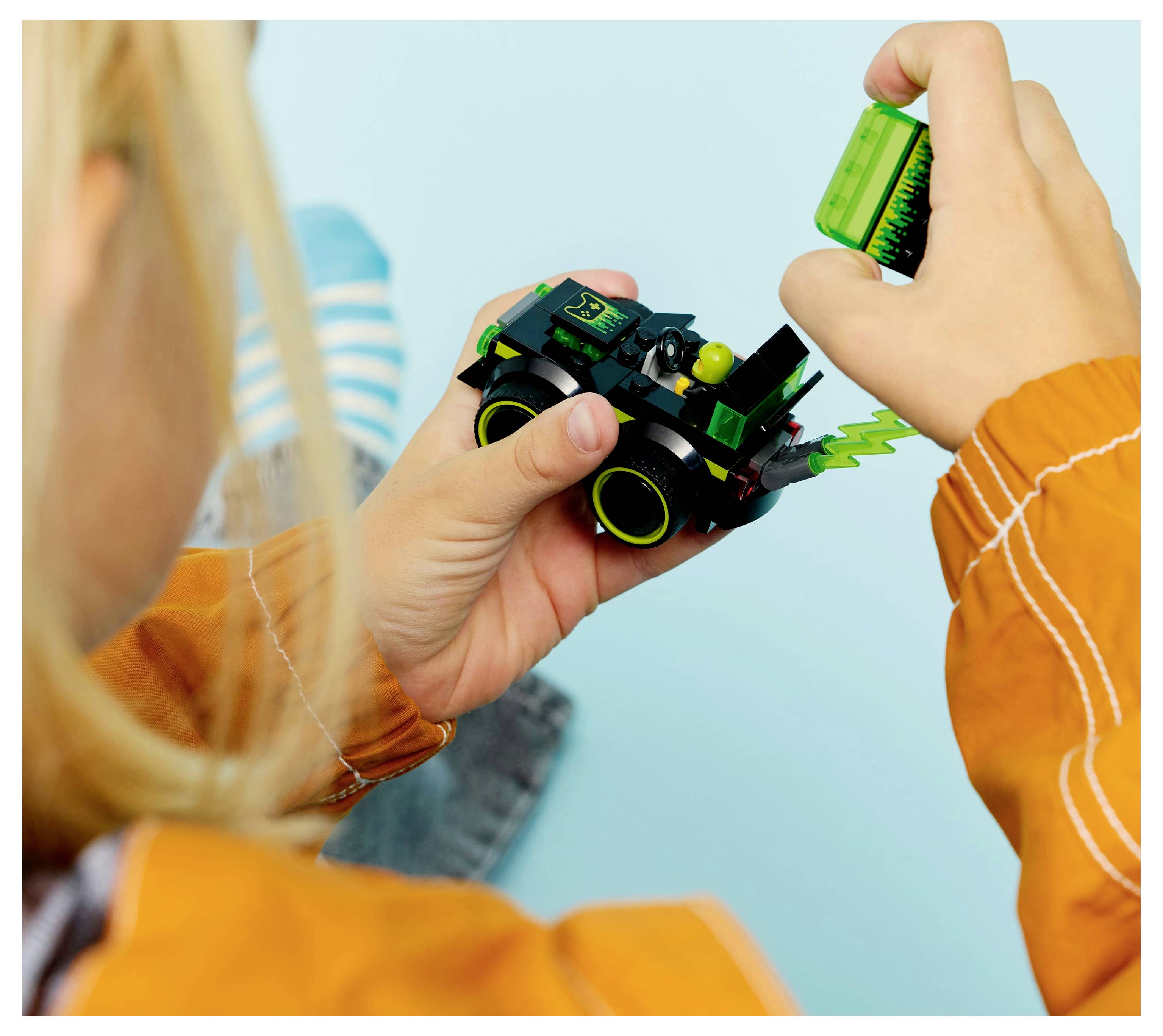 Dítě sestavuje malé hračkové vozidlo, se soustředěným výrazem. Hračka je zelená s žlutými koly a má design blesku. Dětské rukávy jsou jasně oranžové.