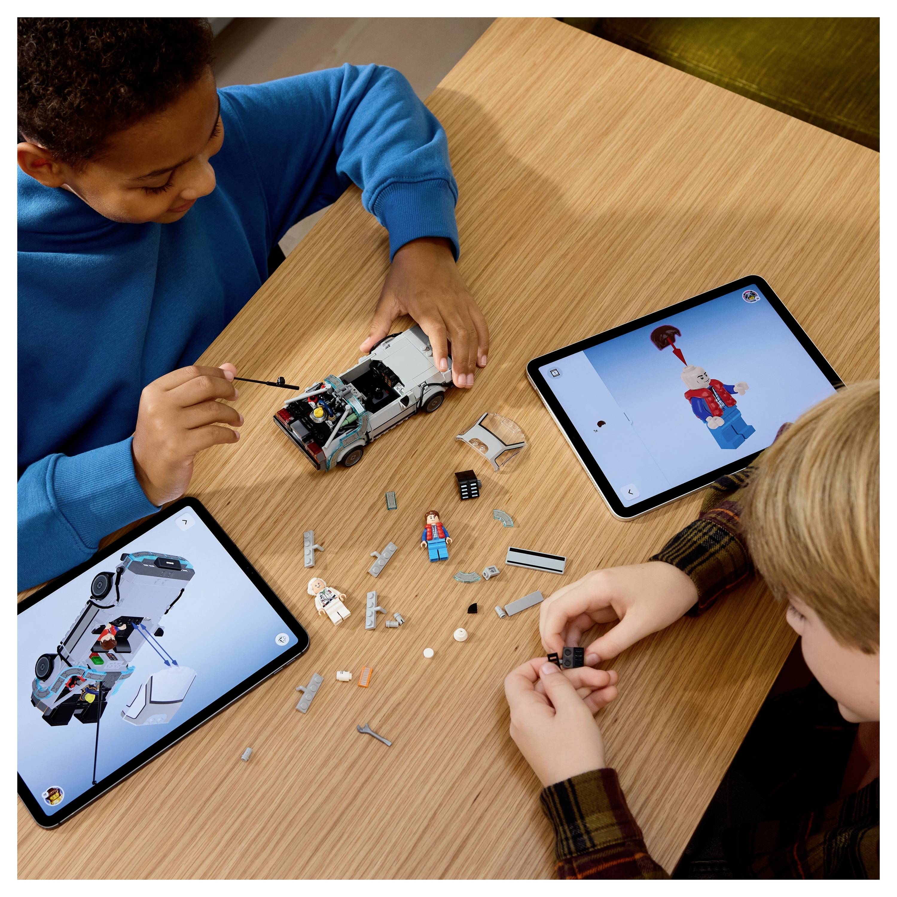Dva děti staví model auta hračky pomocí digitálních instrukcí na tabletech. Díly a částečně sestavené auto leží na stole mezi nimi.