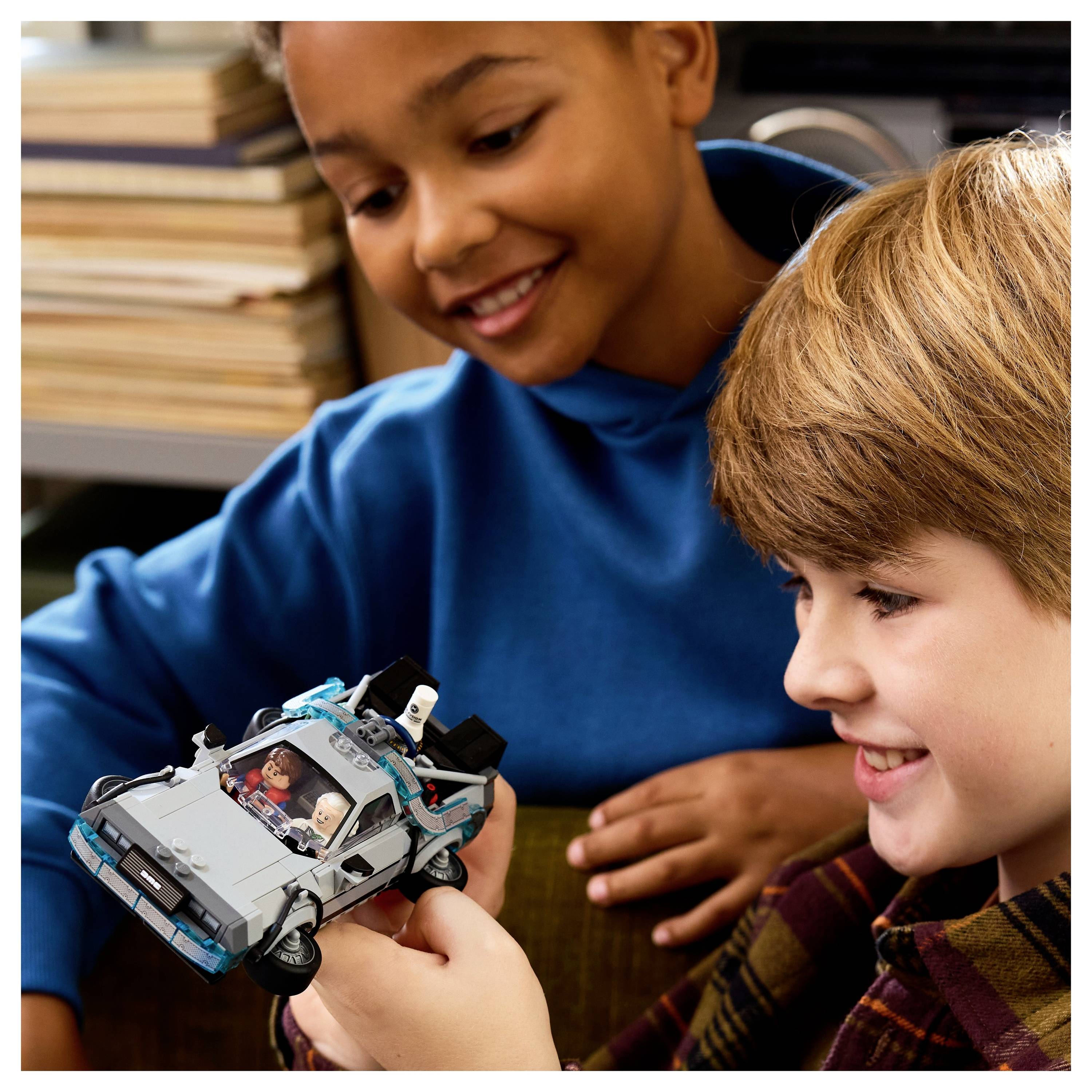 Dvě děti se usmívají a hrají si s hračkářským autem na stole. Jedno dítě drží auto, zatímco druhé je sleduje s nadšením.