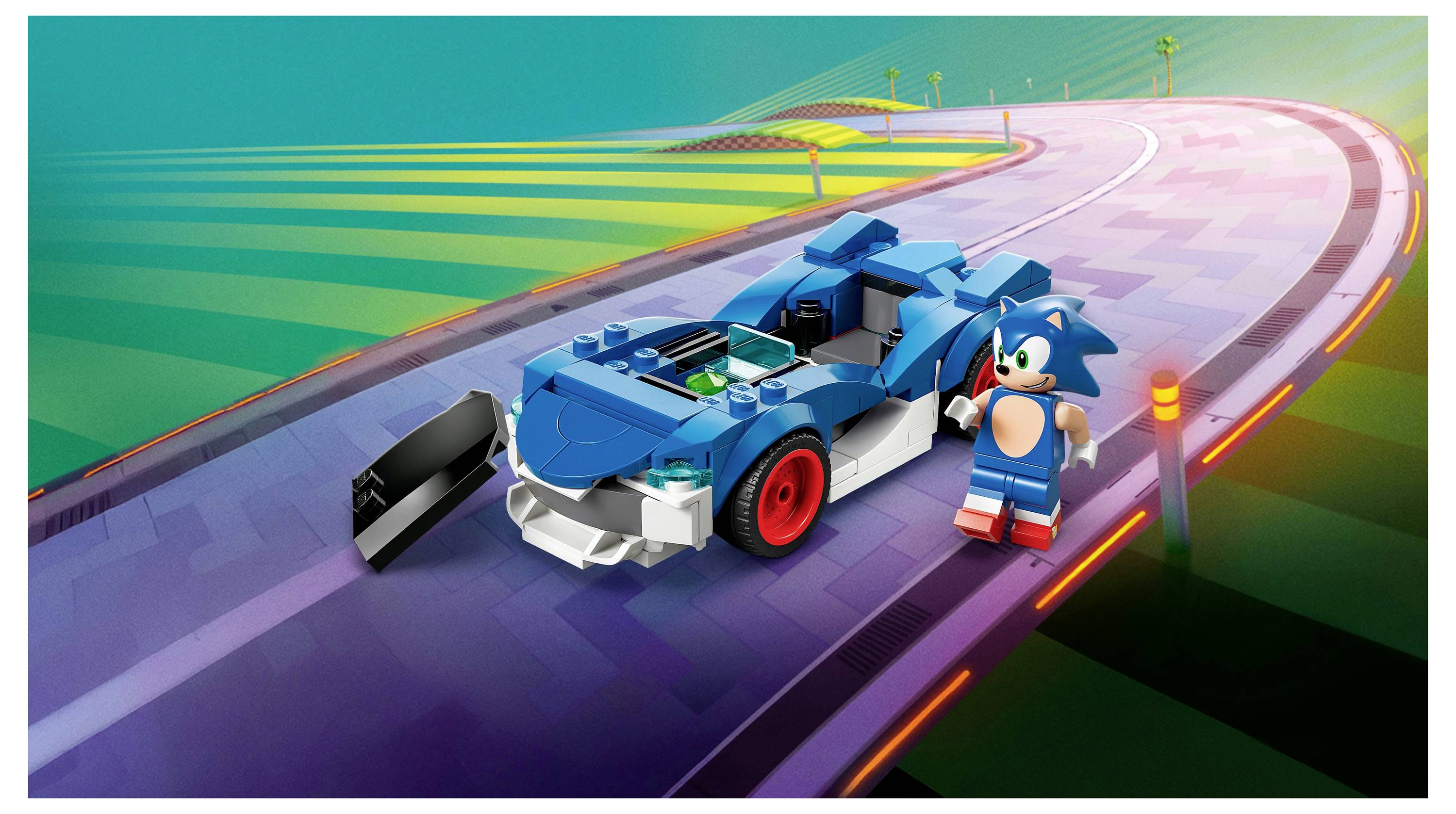 Modrý hračkářský automobil s červenými koly a figurkou podobnou modré ježce stojí na barevné, zakřivené dráze v živém prostředí.