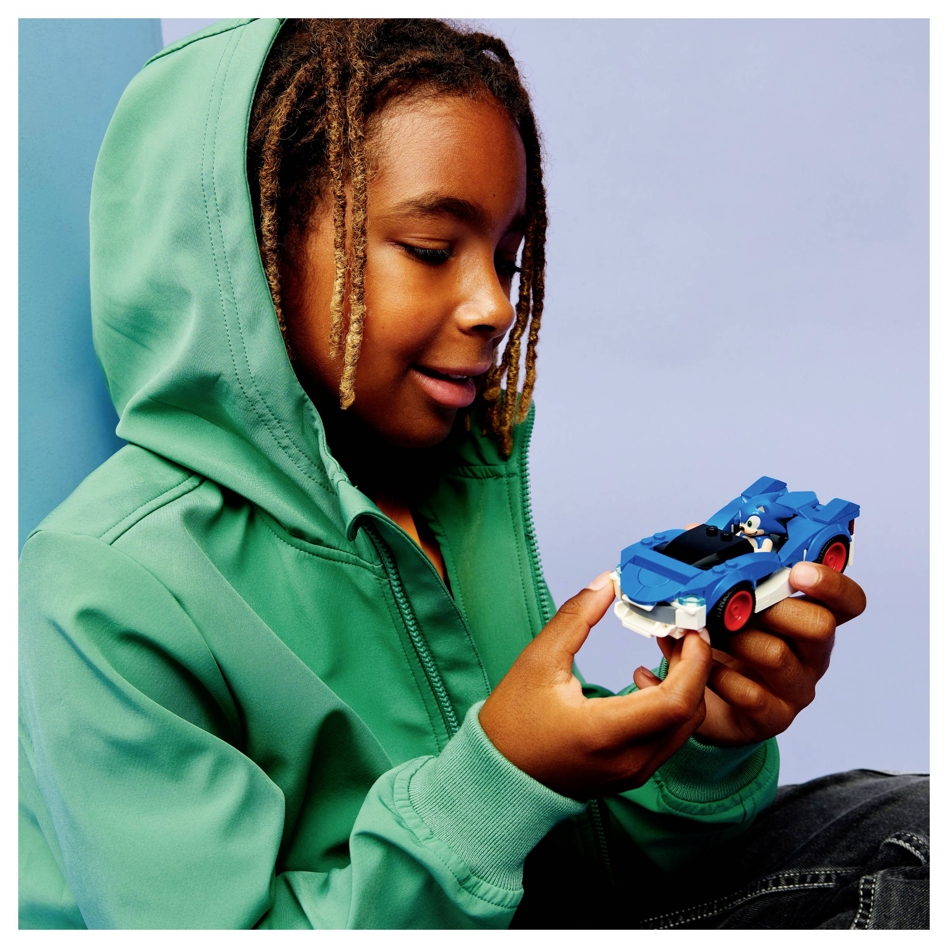 Dítě v zelené mikině se usmívá, zatímco drží malé modré autíčko, sedící před světle modrým pozadím.
