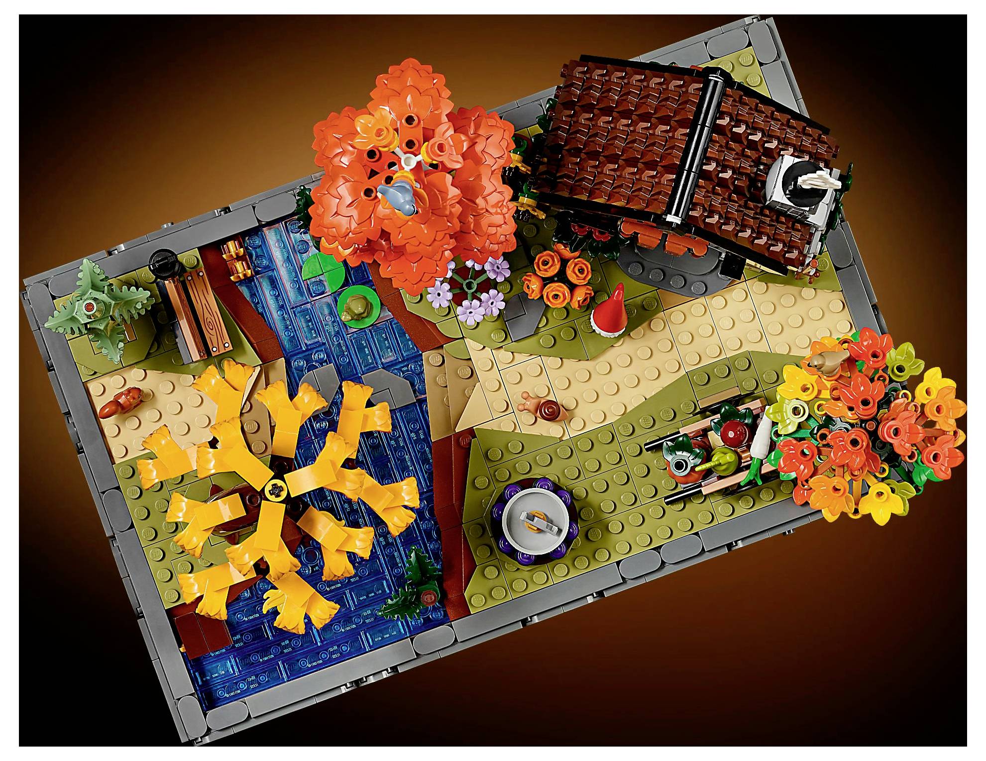 Model krajiny z kostek Lego s podzimními stromy, potokem, cestou a malým cihlových domkem, umístěný na hnědé základně pro podzimní scénu.