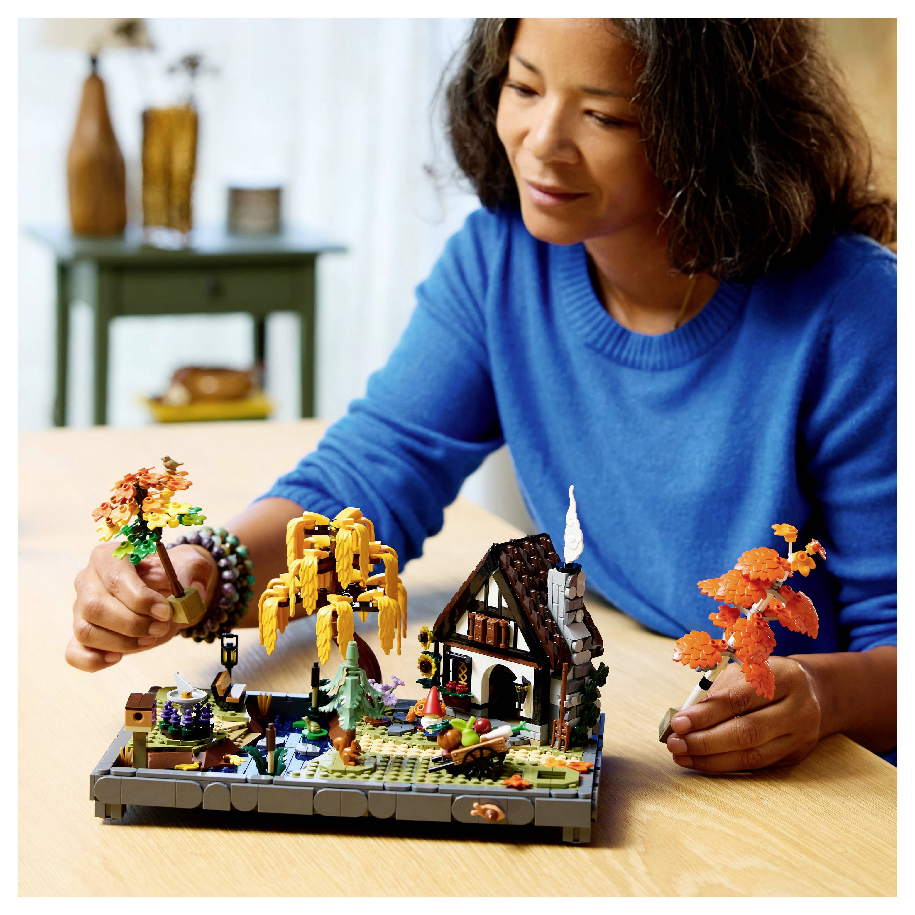 Osoba v modrém svetru staví detailní podzimní LEGO scénu na stole, která zahrnuje stromy a malý dům.