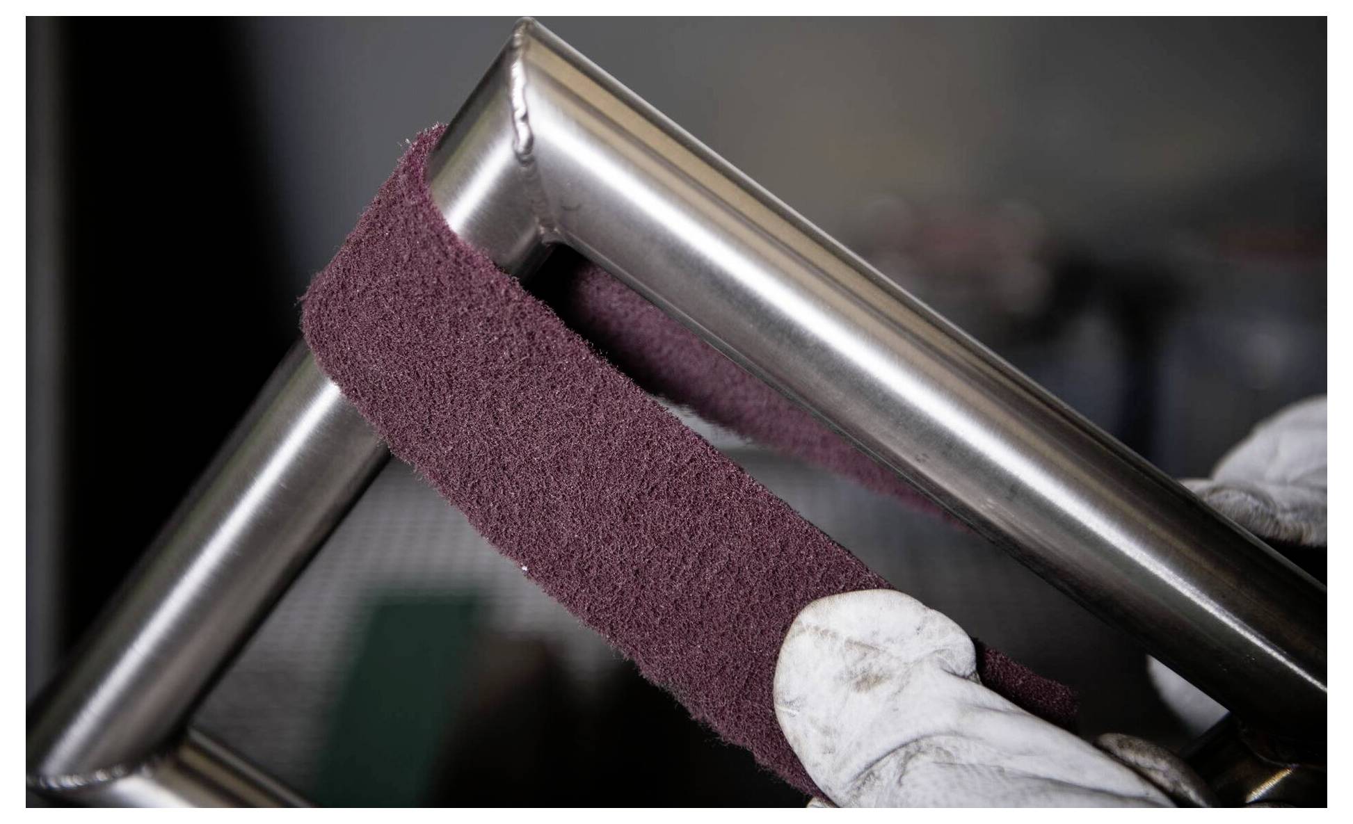 Rukavicemi opatřené ruce leštící nerezový rám pomocí tmavě červeného abrazivního materiálu, přičemž předvádějí proces zjemňování kovových povrchů za účelem dosažení hladkosti.