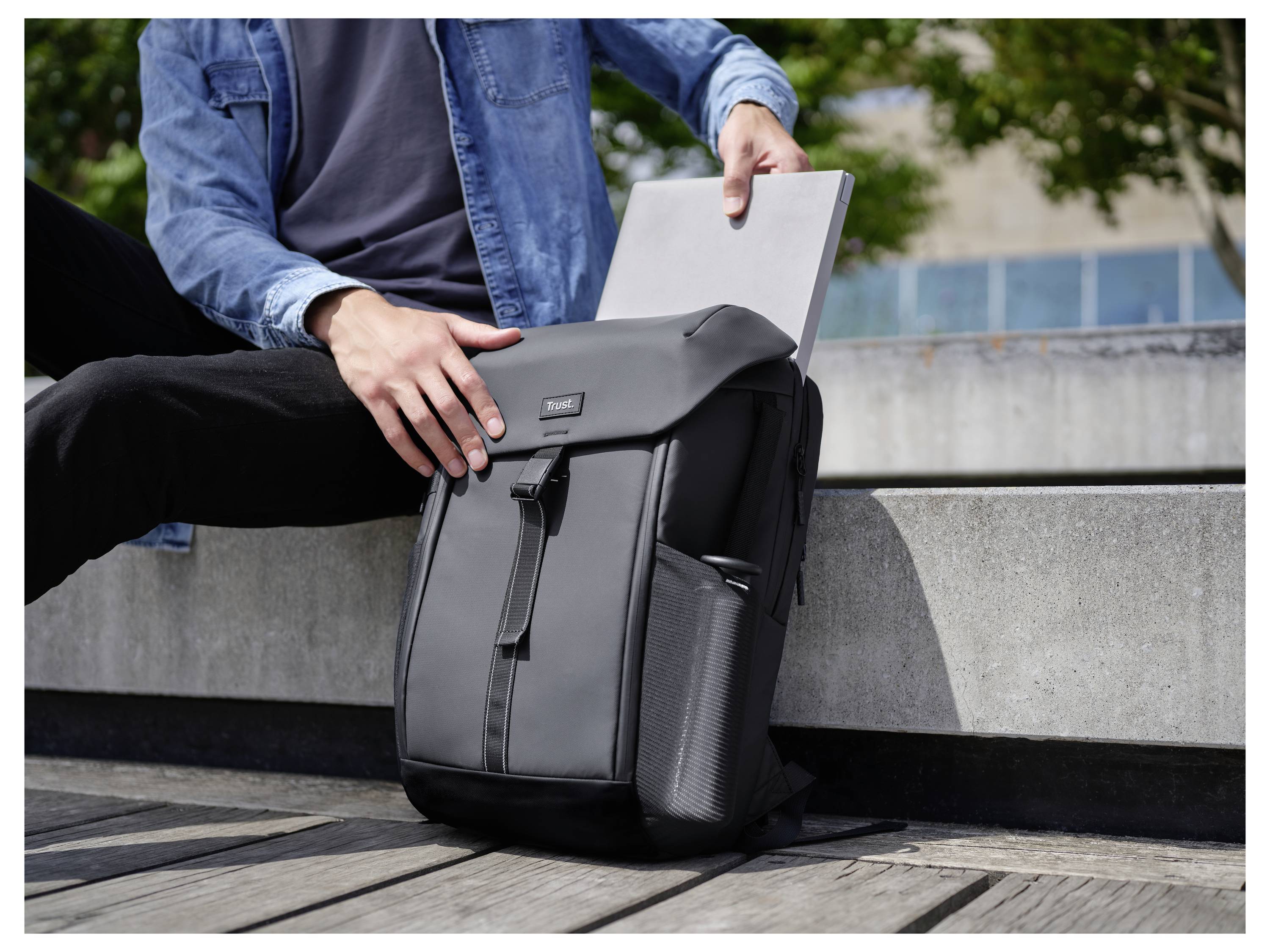 Osoba vkládá laptop do šedého batohu, zatímco sedí venku na betonové římsě.