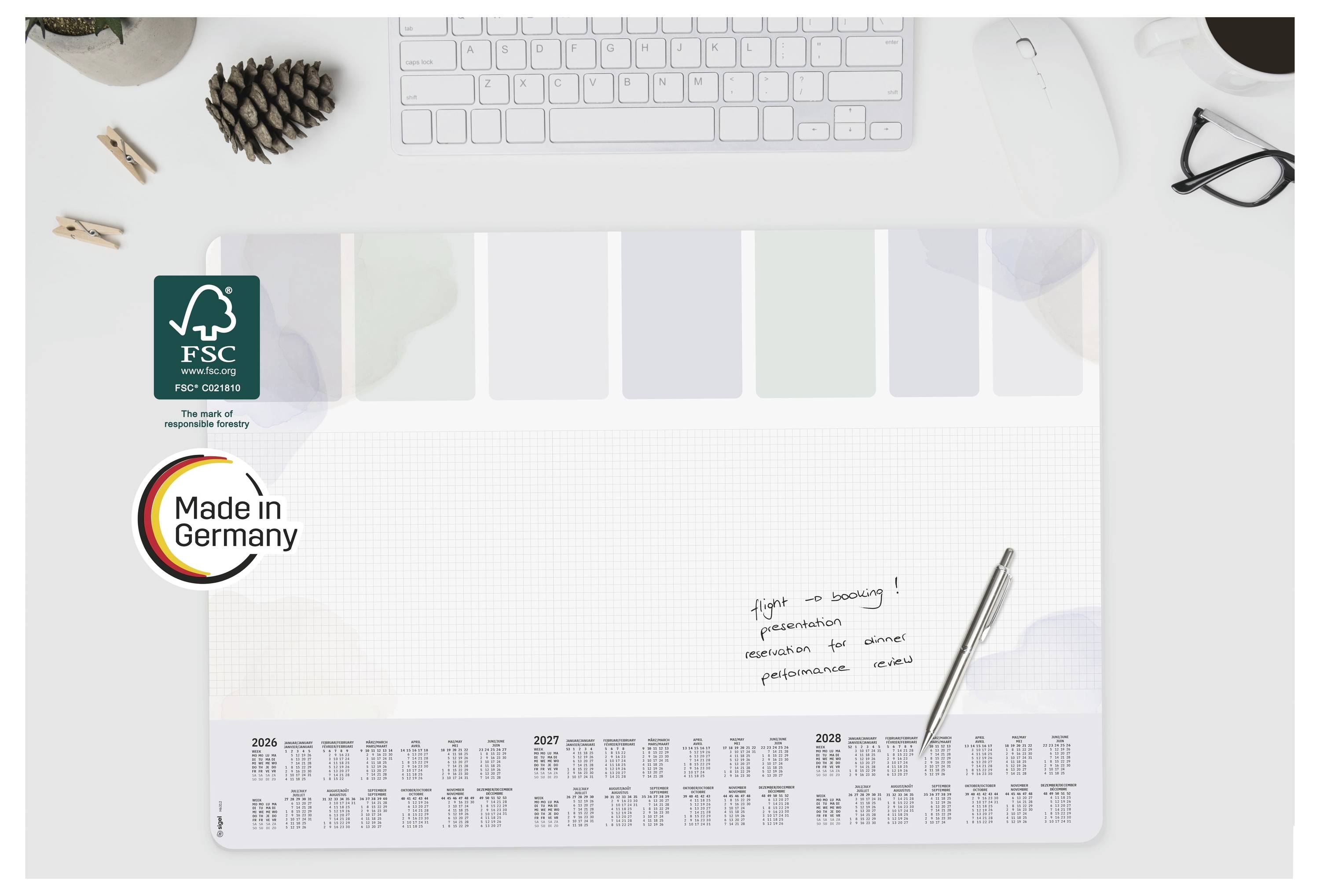 Minimalistický stolní kalendář s certifikacemi FSC a Made in Germany, prázdným centrálním prostorem pro psaní, mřížkou měsíčního plánovače a kancelářskými potřebami kolem něj.