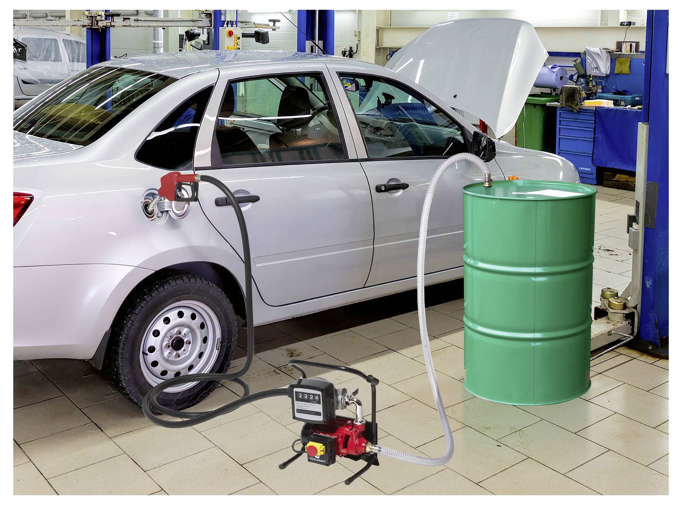Auto v garáži má hadici připojenou z palivové nádrže k zelené barely. Kapota vozu je otevřená a v pozadí jsou viditelné nástroje.