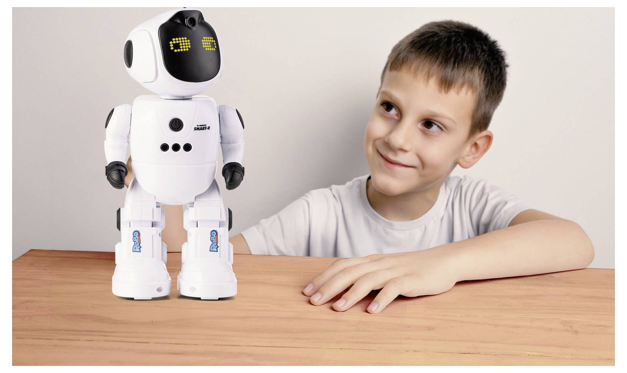 Mladý chlapec se usmívá na malého bílého robota s digitálníma očima sedícího na stole, což naznačuje interakci mezi dítětem a technologií.