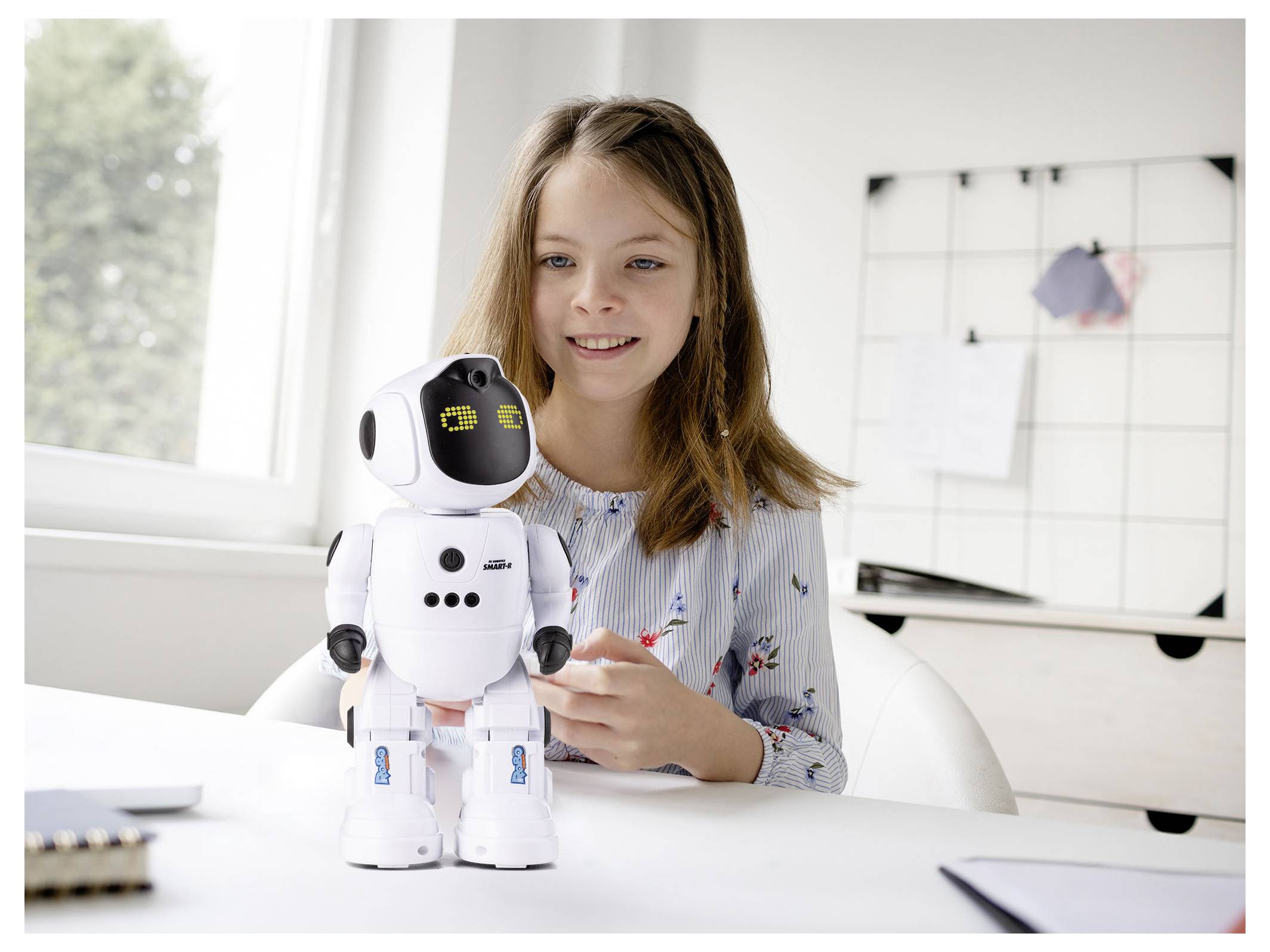 Mladá dívka sedí u stolu a usmívá se na malého bílého robota stojícího před ní, což naznačuje interakci nebo studium doma.