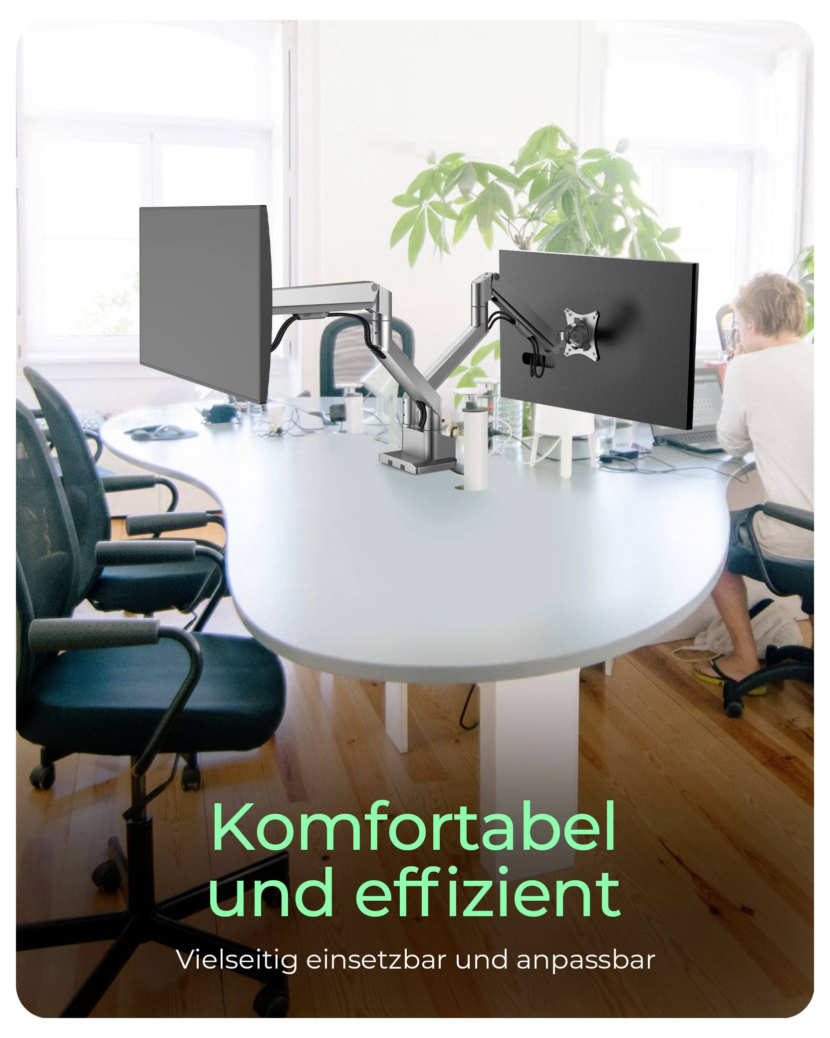 Dva počítačové monitory na ergonomickém rameni nad kulatým kancelářským stolem. V pozadí někdo píše na laptopku.