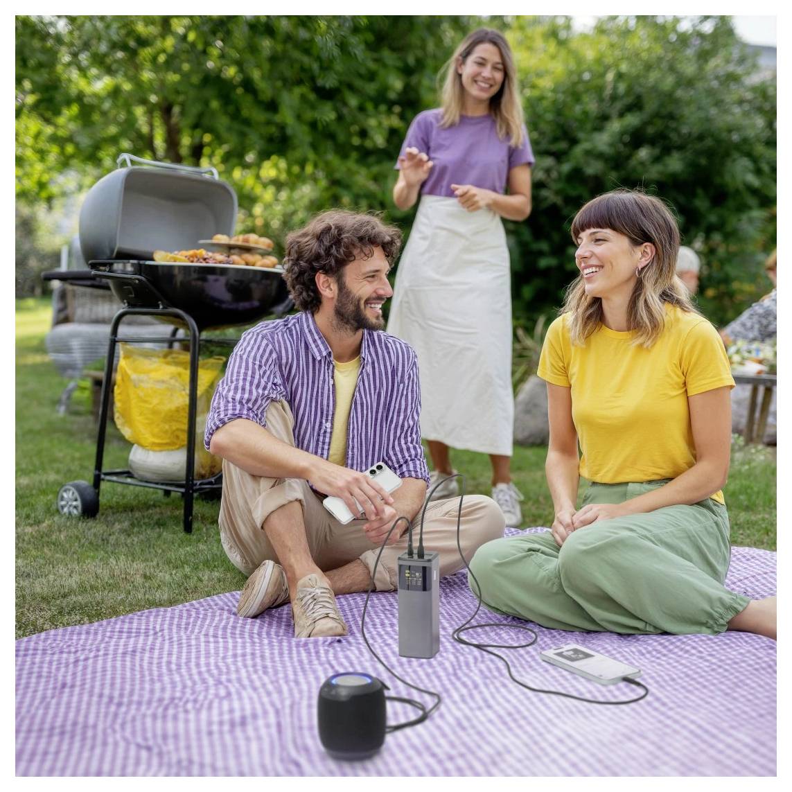 Muž a žena sedí na piknikovém přikrytí, usmívají se a povídají si. V pozadí je gril na barbecue a další žena stojí poblíž.