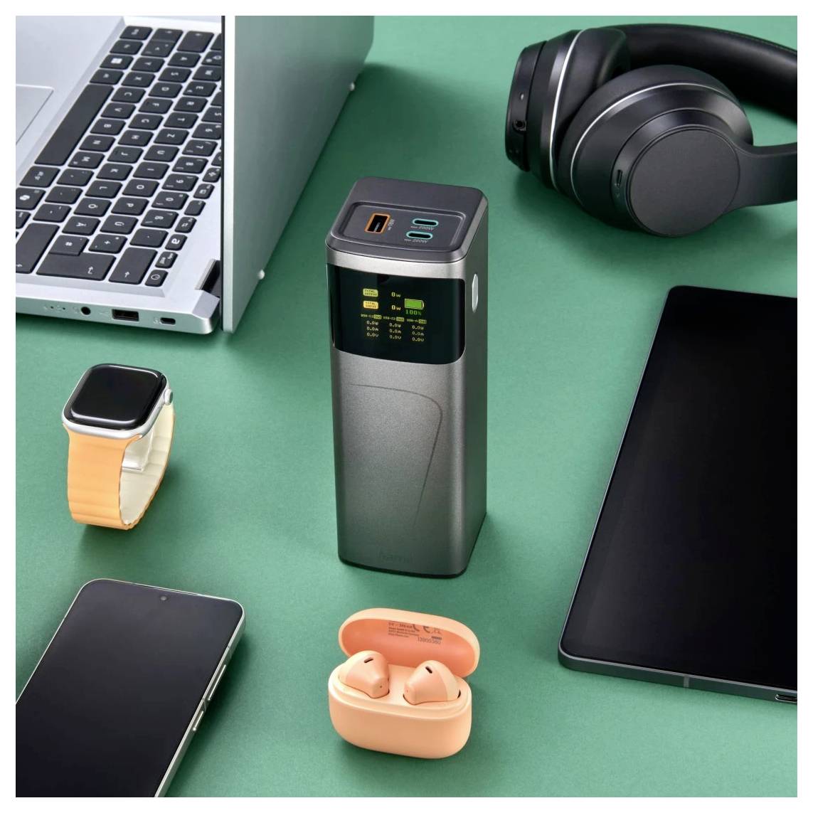Desktopové uspořádání s uzavřeným laptopem, sluchátky, chytrými hodinkami, smartphonem, tabletem, oranžovými peckami do uší a přenosným nabíjecím zařízením na zelené ploše.