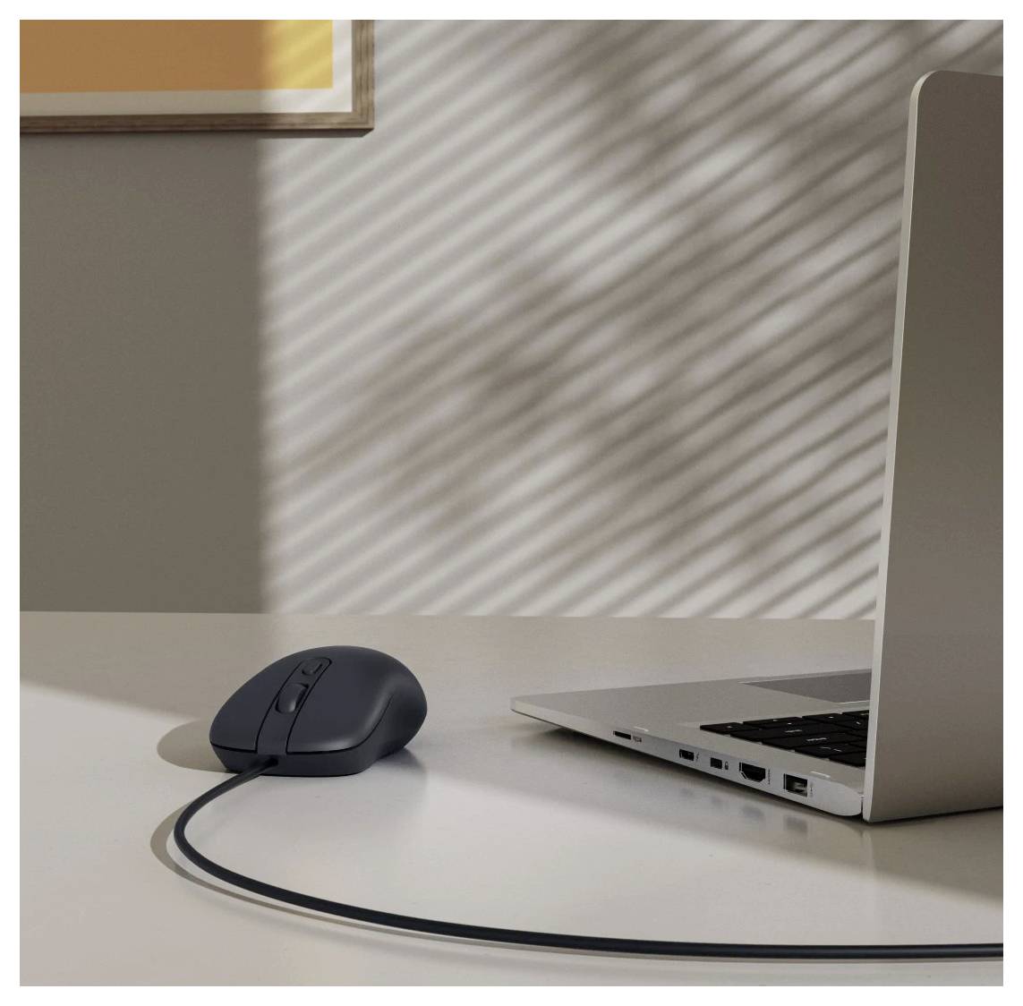 Elegantní černá počítačová myš připojená k stříbrnému laptopu na bílém stole, přičemž sluneční světlo vrhá pruhované stíny skrz okenní žaluzii.