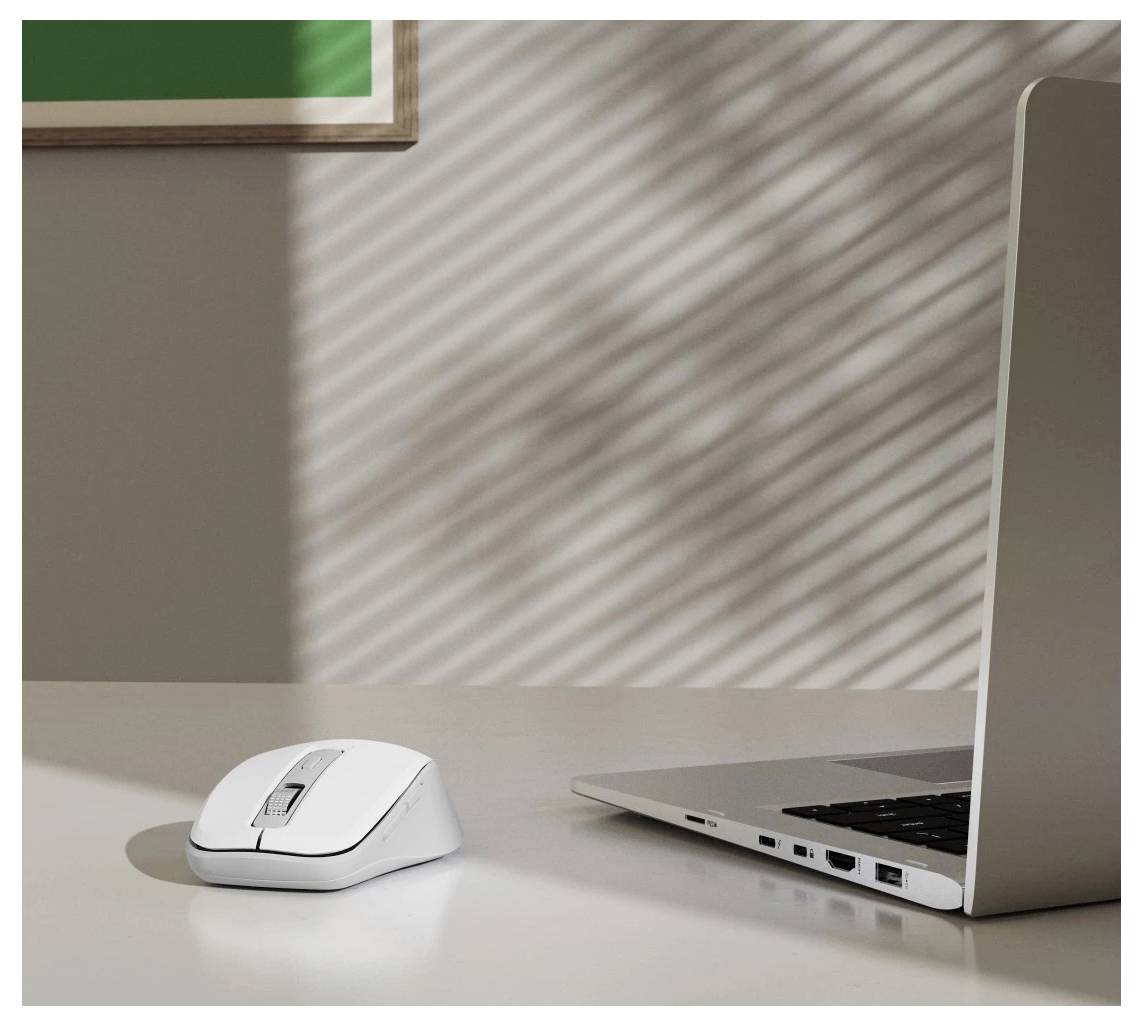 Bílá bezdrátová myš vedle stříbrného laptopu na stole. Sluneční světlo vytváří stínové vzory žaluzií na stěně a povrchu stolu.