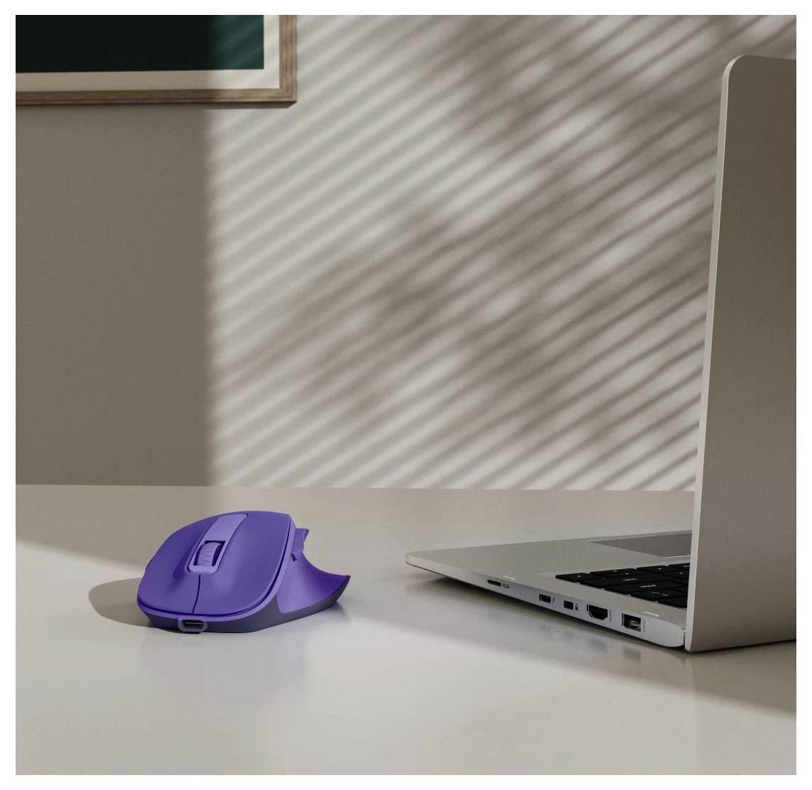 Fialová počítačová myš leží na stole vedle otevřeného laptopů, přičemž sluneční světlo vrhá pruhované stíny přes povrch.