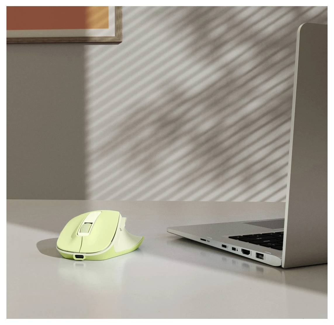 Zelená bezdrátová počítačová myš položená vedle otevřeného laptopu na stole, přičemž sluneční světlo vrhá pruhované stíny skrze žaluzie.