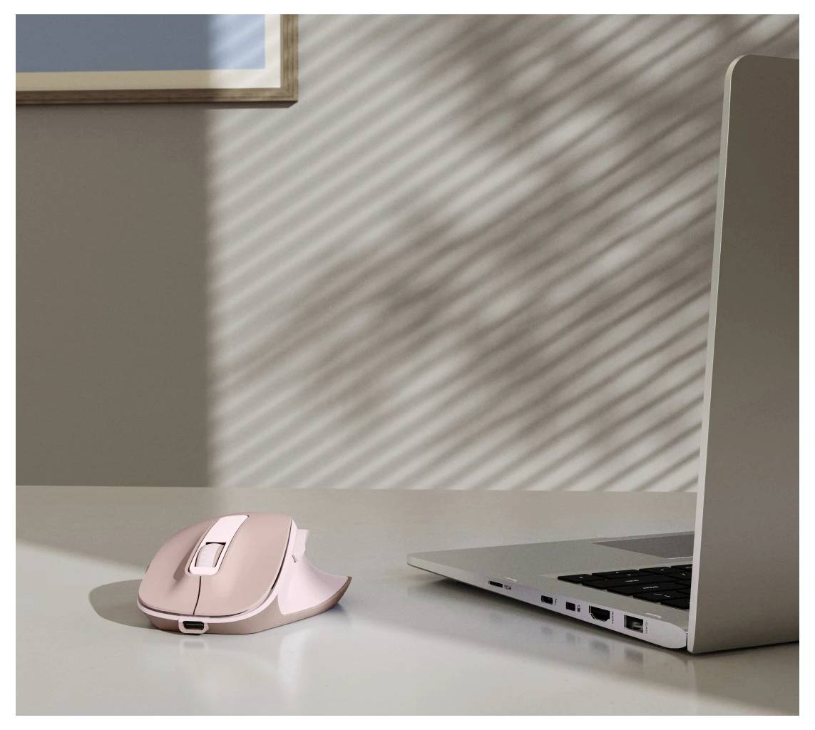 Růžová ergonomická bezdrátová myš na bílém stole vedle otevřeného stříbrného laptopu, s slunečním světlem vrhajícím pruhované stíny skrze okno.