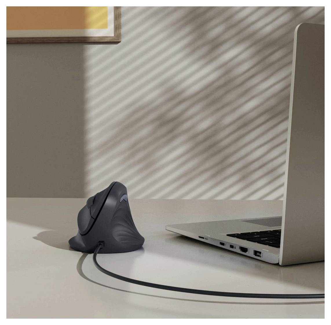 Vertikální ergonomická myš připojená k laptopu leží na stole. Sluneční světlo vrhá stíny skrz žaluzie a vytváří pruhovaný vzor na zdi.