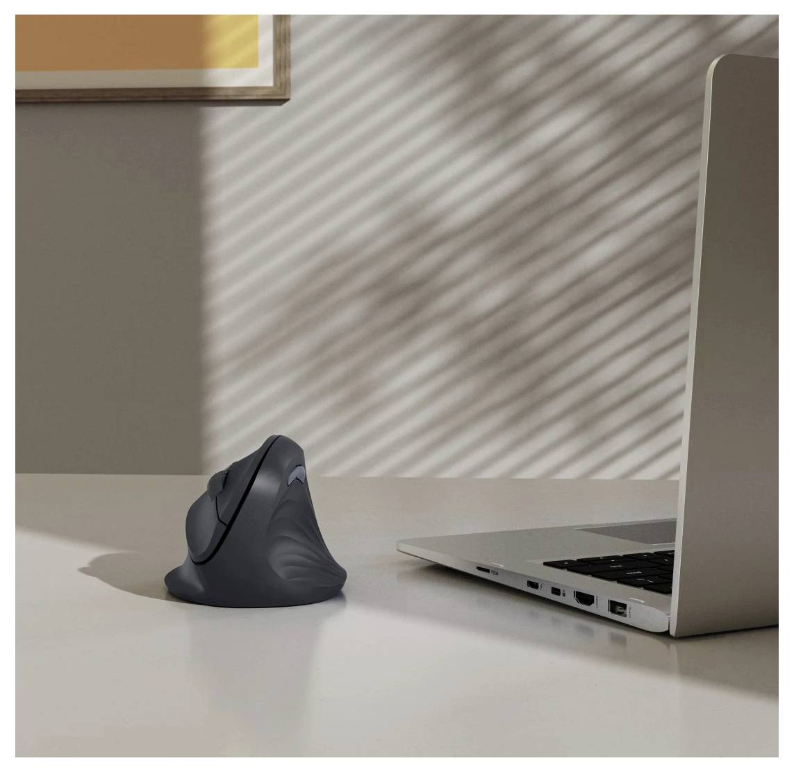 Vertikální ergonomická myš vedle otevřeného laptopu na sluncem osvětleném stole, která naznačuje pracoviště zaměřené na pohodlí a produktivitu.