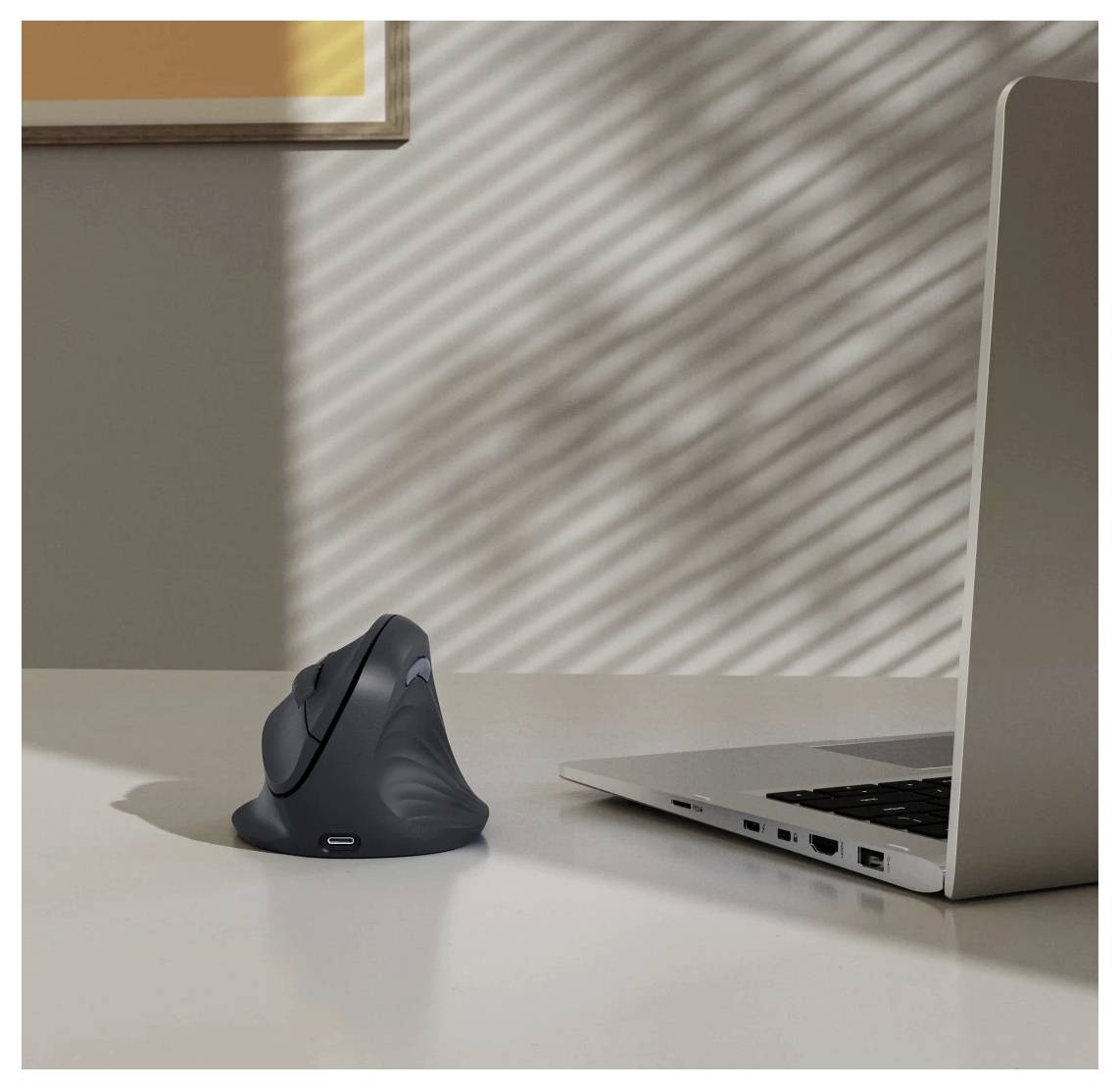 Vertikální bezdrátová ergonomická myš umístěná vedle laptopu na stole, s pruhovanými slunečními vzory na stěně a pracovní desce.