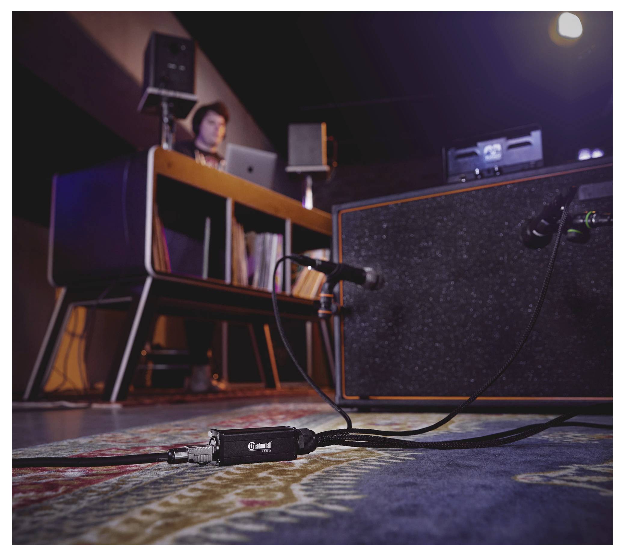 Nahrávací studio zaměřené na kytarový efekt a kabel ležící na koberci, propojený s hudebním vybavením. V pozadí je vidět osoba sedící u stolu s audiotechnikou.
