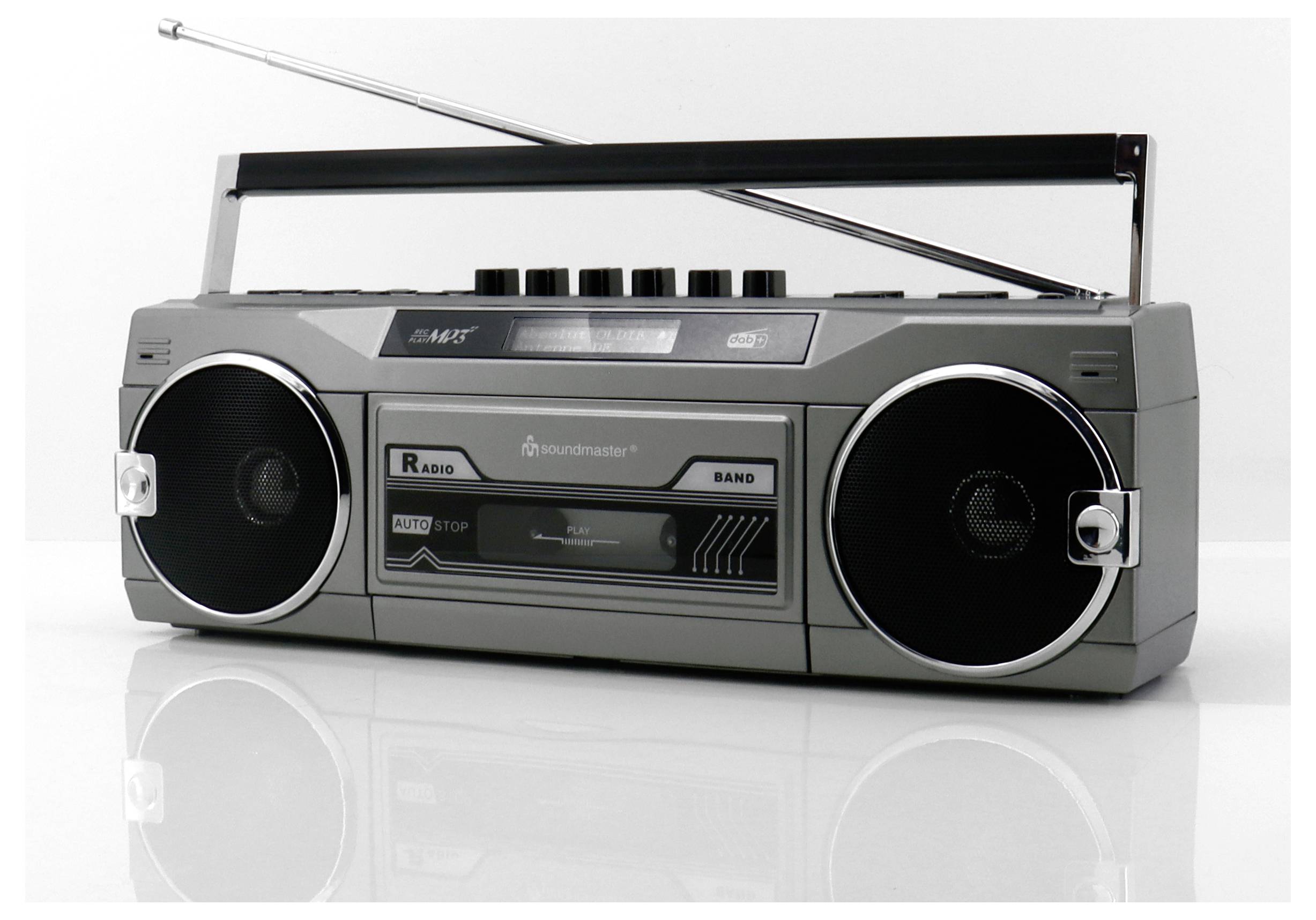 Vintage šedý radiomagnetofon s vysunutou anténou a dvěma velkými reproduktory po stranách, umístěný na lesklém bílém povrchu.