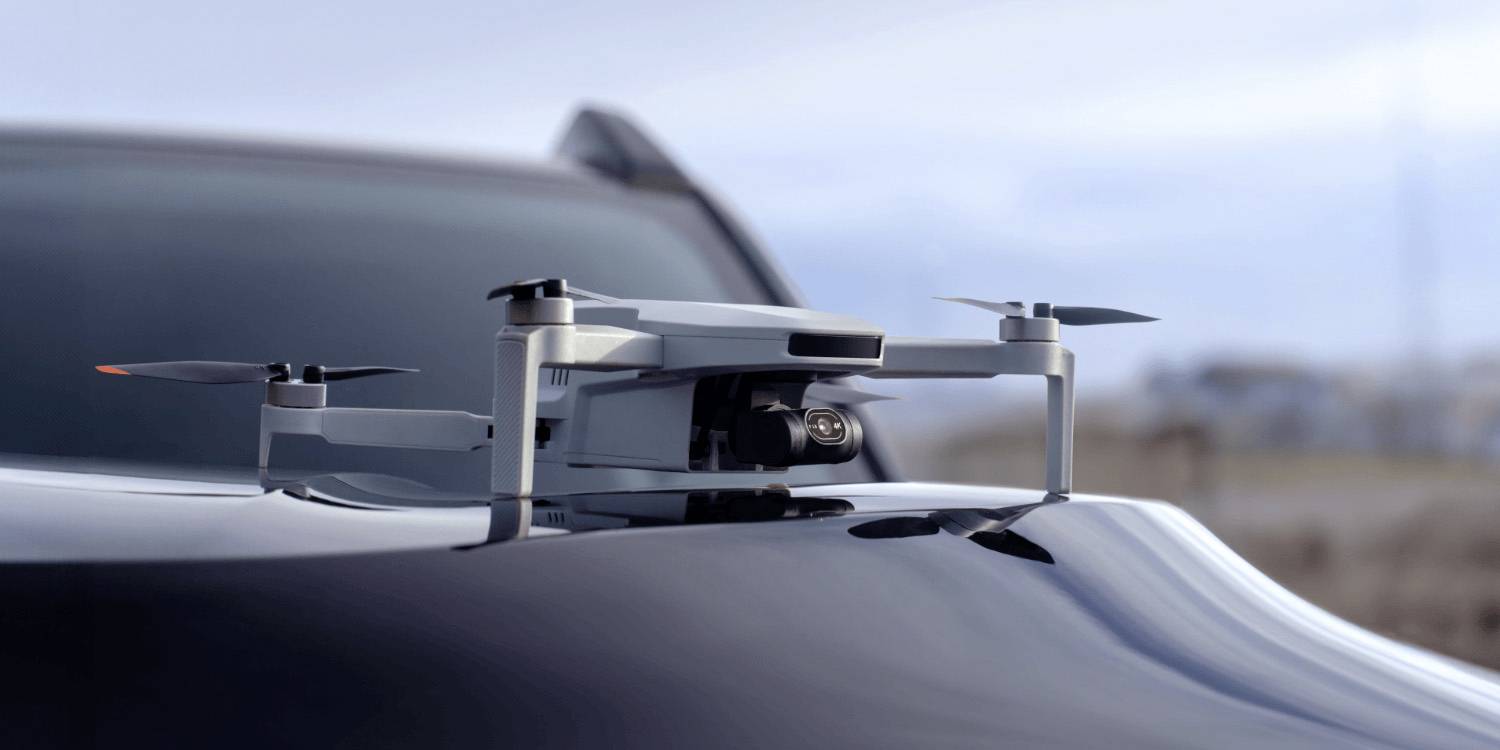 Dron odpočívá na kapotě elegantního vozu v rozmazaném venkovním prostředí, což naznačuje témata pokročilé technologie nebo dopravy.
