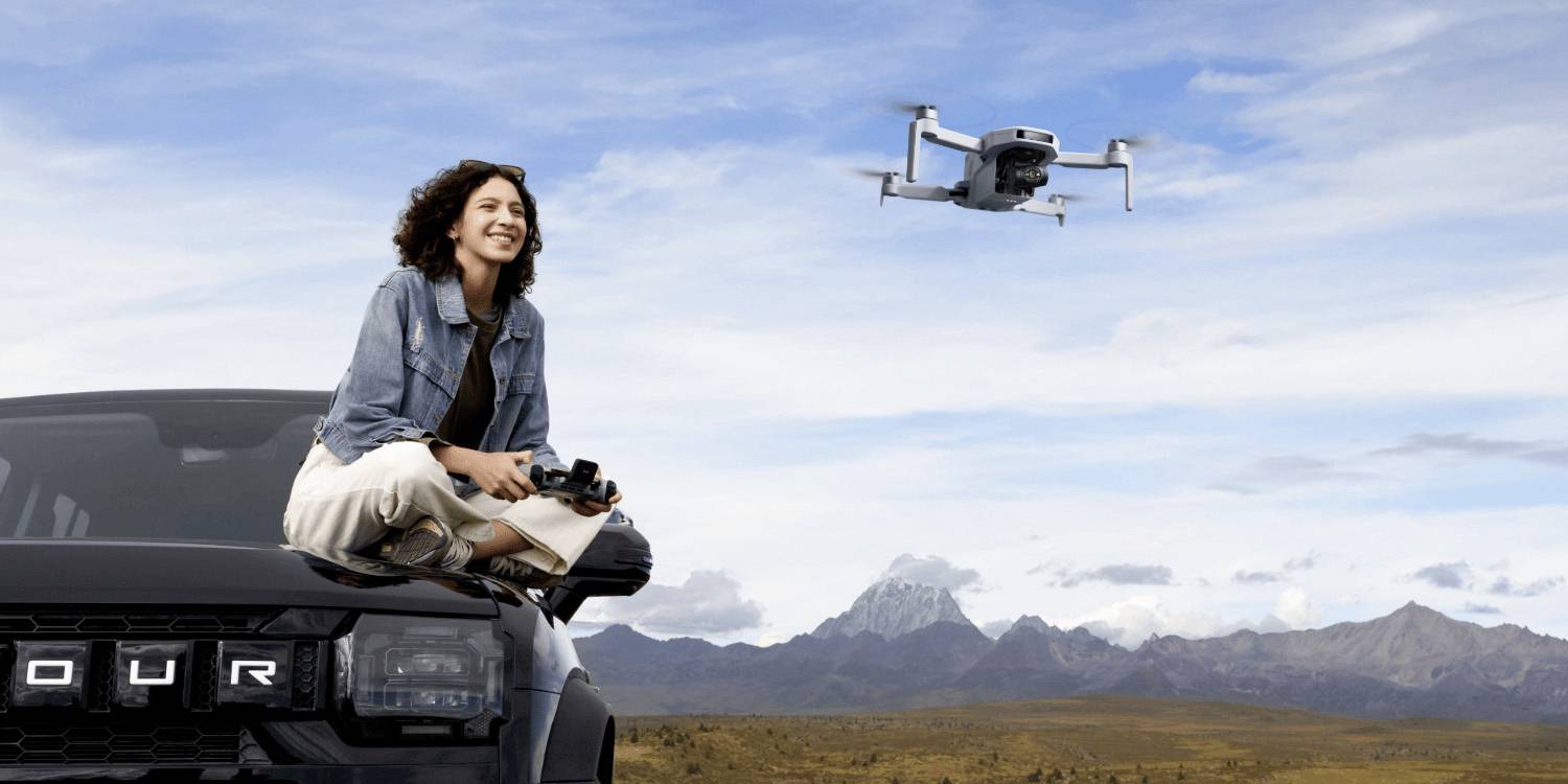 Žena sedí na černém SUV 4x4 a drží dálkové ovládání dronu. Poblíž létá dron nad scenickou krajinou s horami v pozadí.