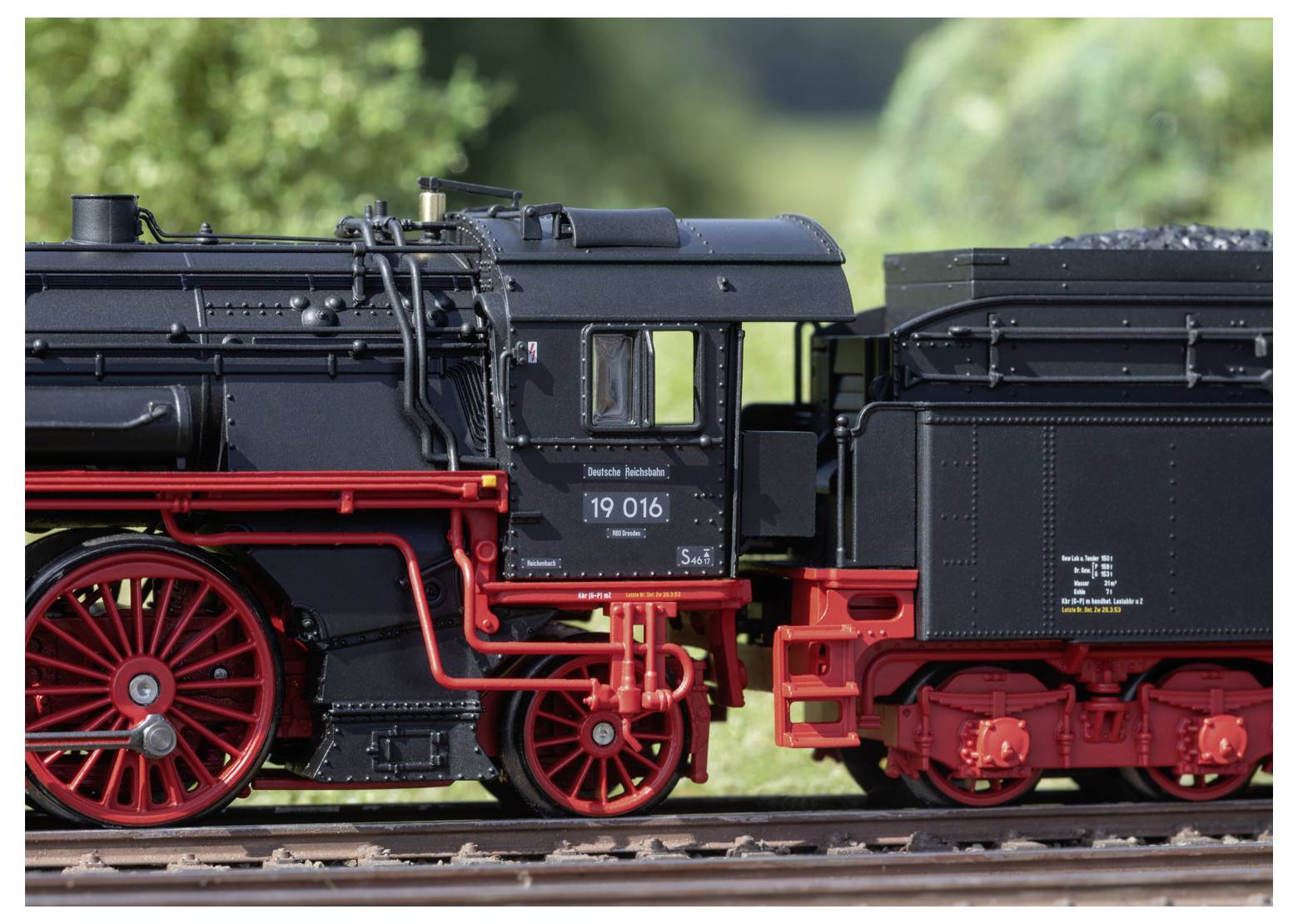 Detailní model parní lokomotivy s červenými koly a černým tělem, zobrazující složité mechanické detaily, umístěný před rozmazaným zeleným pozadím.