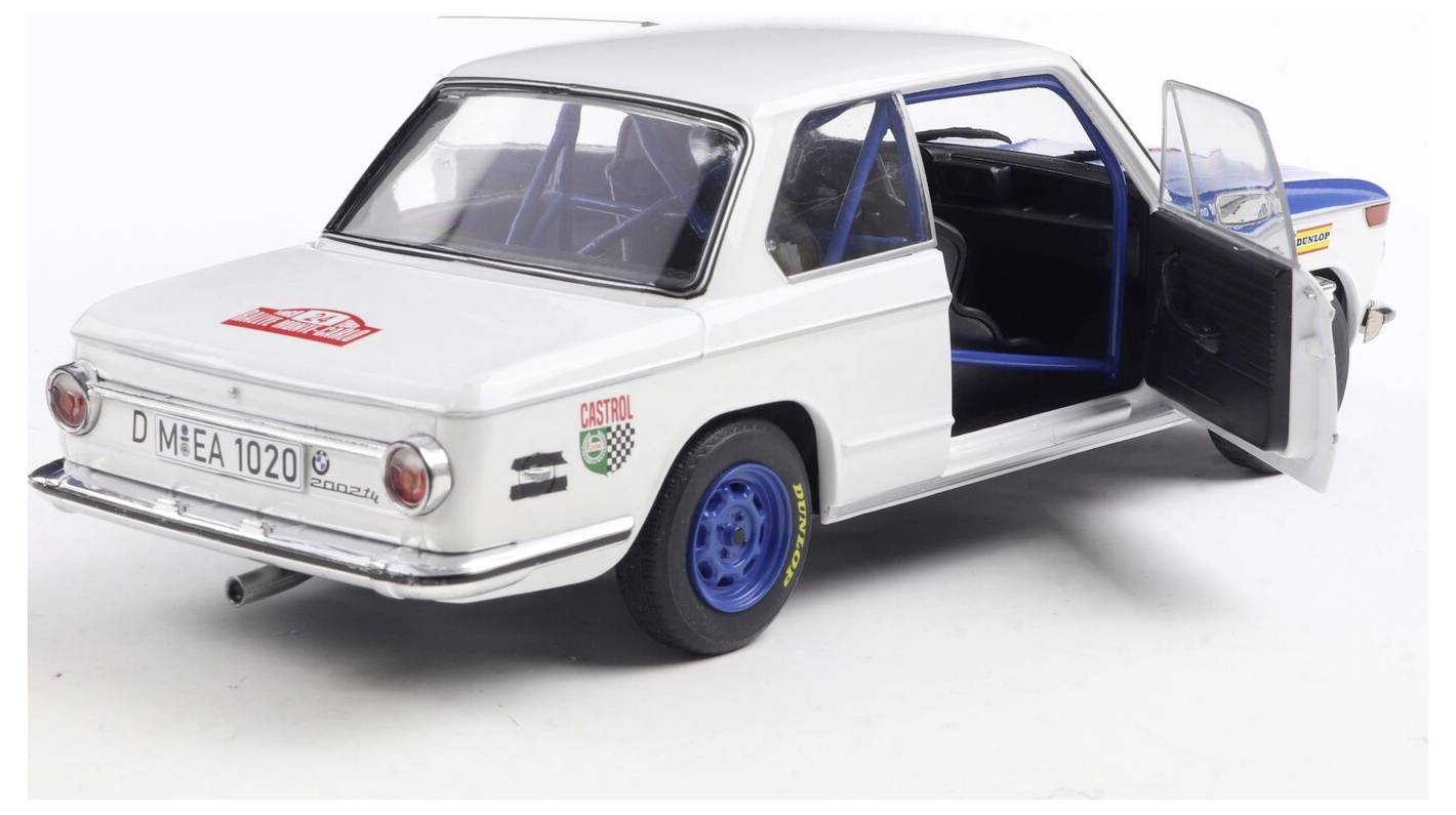 Bílý vintage model závodního automobilu s modrými koly, otevřenými dveřmi a závodními samolepkami na boku a zadní části, umístěný na jednoduchém pozadí.