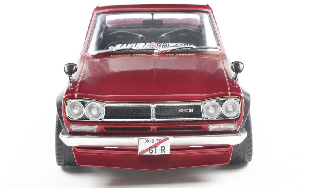 'Pohled zepředu na červené vintage auto s klasickým designem. Na poznávací značce je napsáno 'GT-R' a na čelním skle je viditelný nápis 'SAKURA'.'